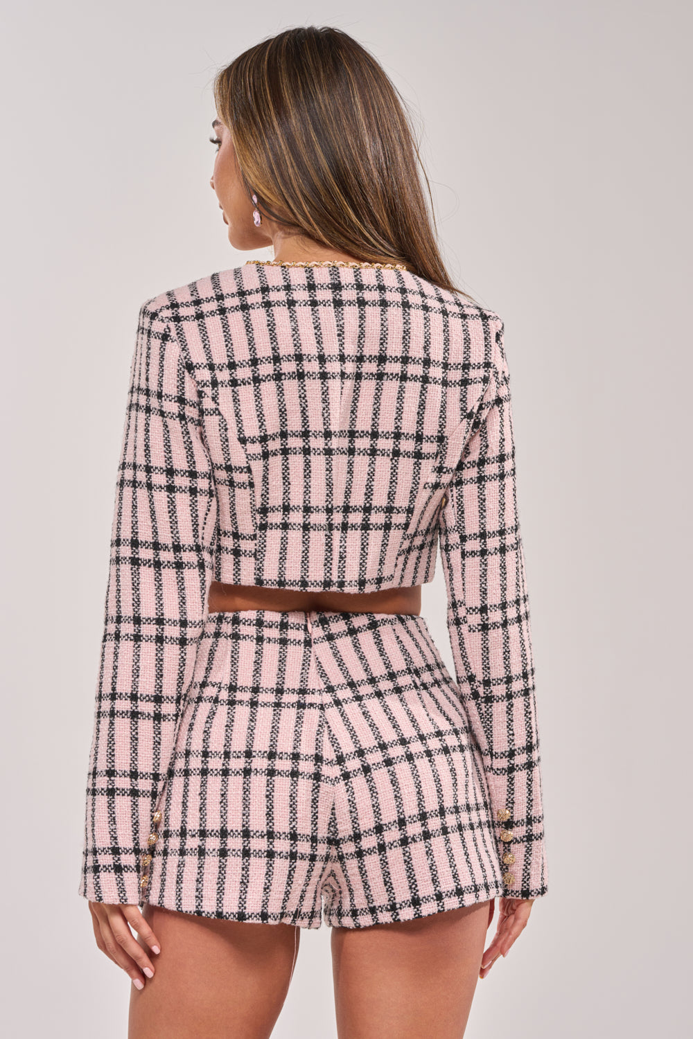 A woman with straight, brown hair stands facing away, wearing the LOOK AT ME BABY TWEED JACKET—a pink plaid cropped style with gold hardware—paired with matching high-waisted shorts. The background is plain and light-colored. 5