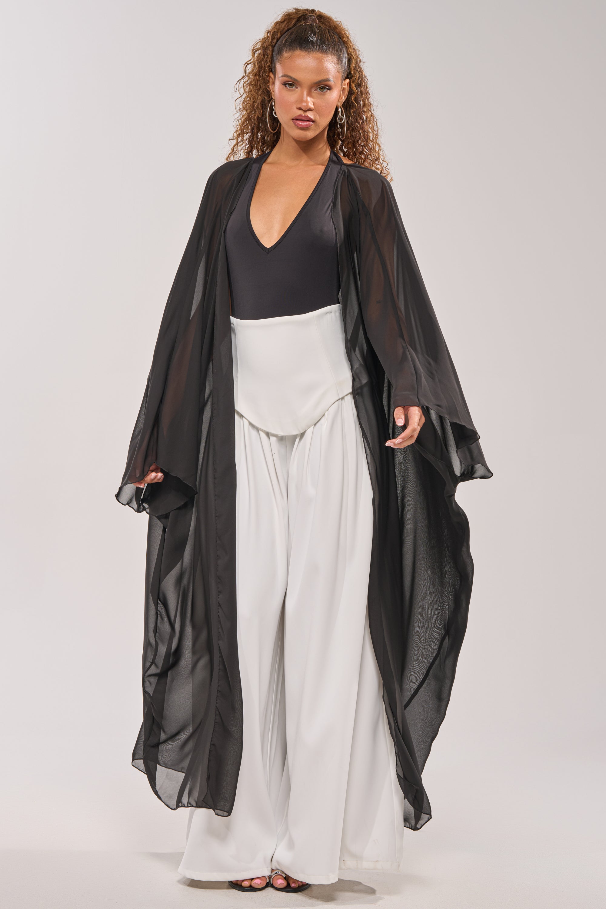 A woman poses in the ADDAMS CAPE BODYSUIT, styled with a sheer black kimono and wide-leg white pants, creating a striking editorial look against a plain light background. 0