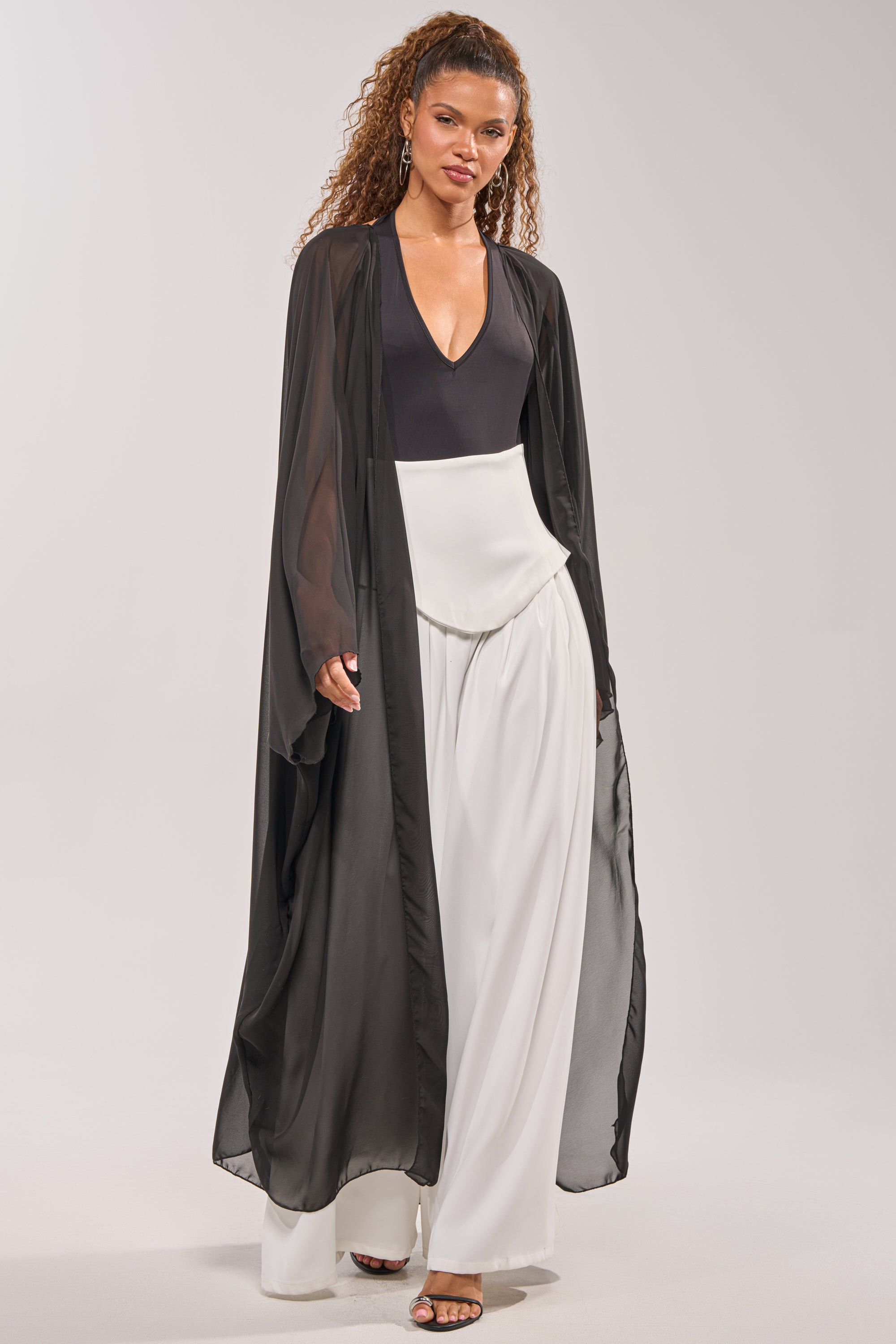A woman stands confidently facing forward with her hair in loose curls, wearing the ADDAMS CAPE BODYSUIT and wide-leg white pants, topped with strappy heels and a sheer black long duster. 1