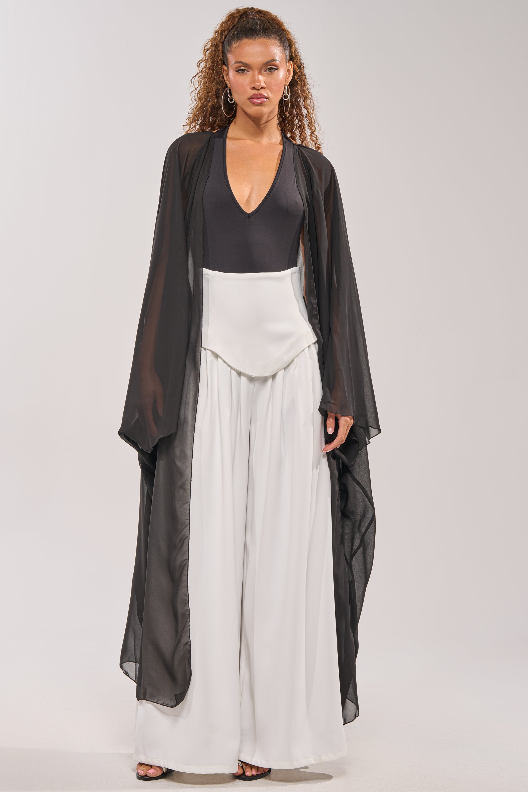 A woman with curly hair wears the ADDAMS CAPE BODYSUIT under a sheer long black robe and high-waisted, wide-leg white pants. She faces forward against a light background, creating an editorial look. 2