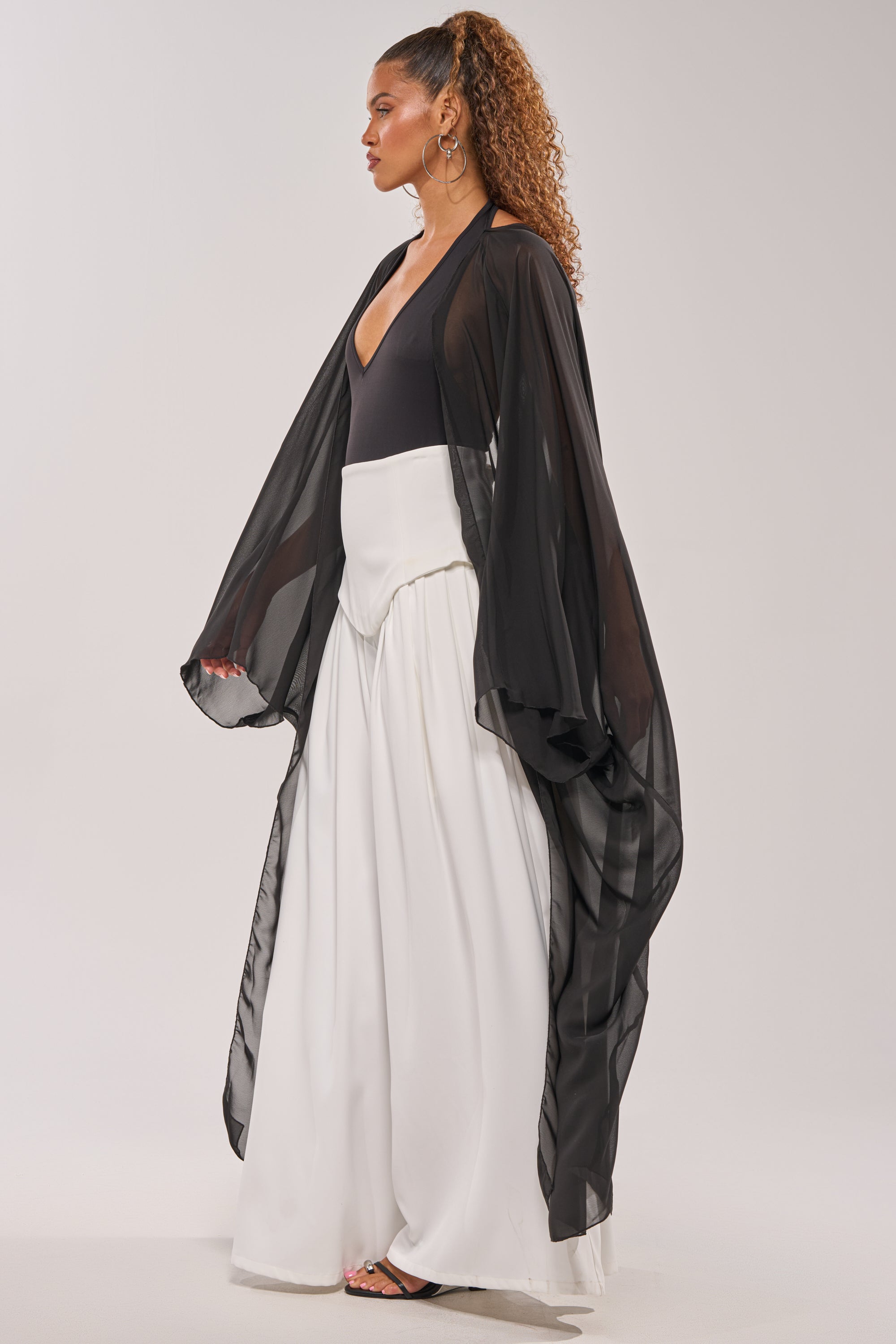 A woman stands in profile wearing the ADDAMS CAPE BODYSUIT styled with a sheer black kimono robe, flowing white pants, and black sandals. Her curly hair is in a high ponytail as she poses against a plain light background. 3