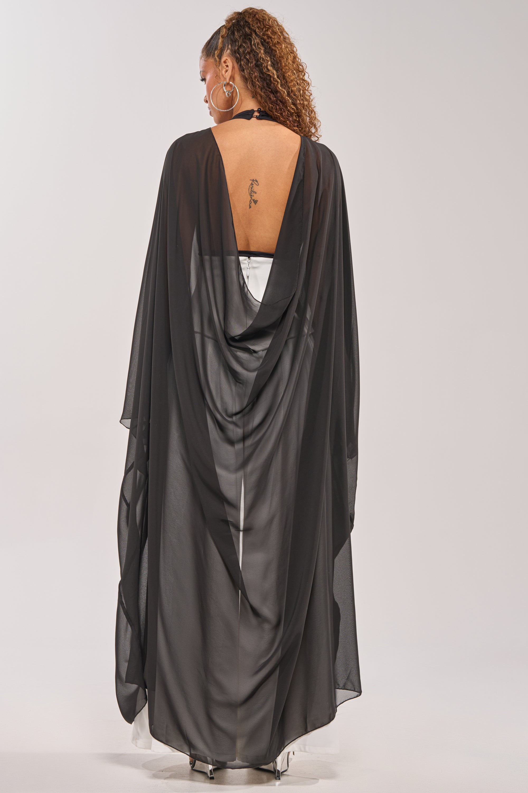 A woman with curly hair and hoop earrings models the ADDAMS CAPE BODYSUIT—a black halter cape style, open back revealing a tattoo, layered over white. The deep neckline adds statement flair, ideal for special occasions. She stands before a plain backdrop. 4