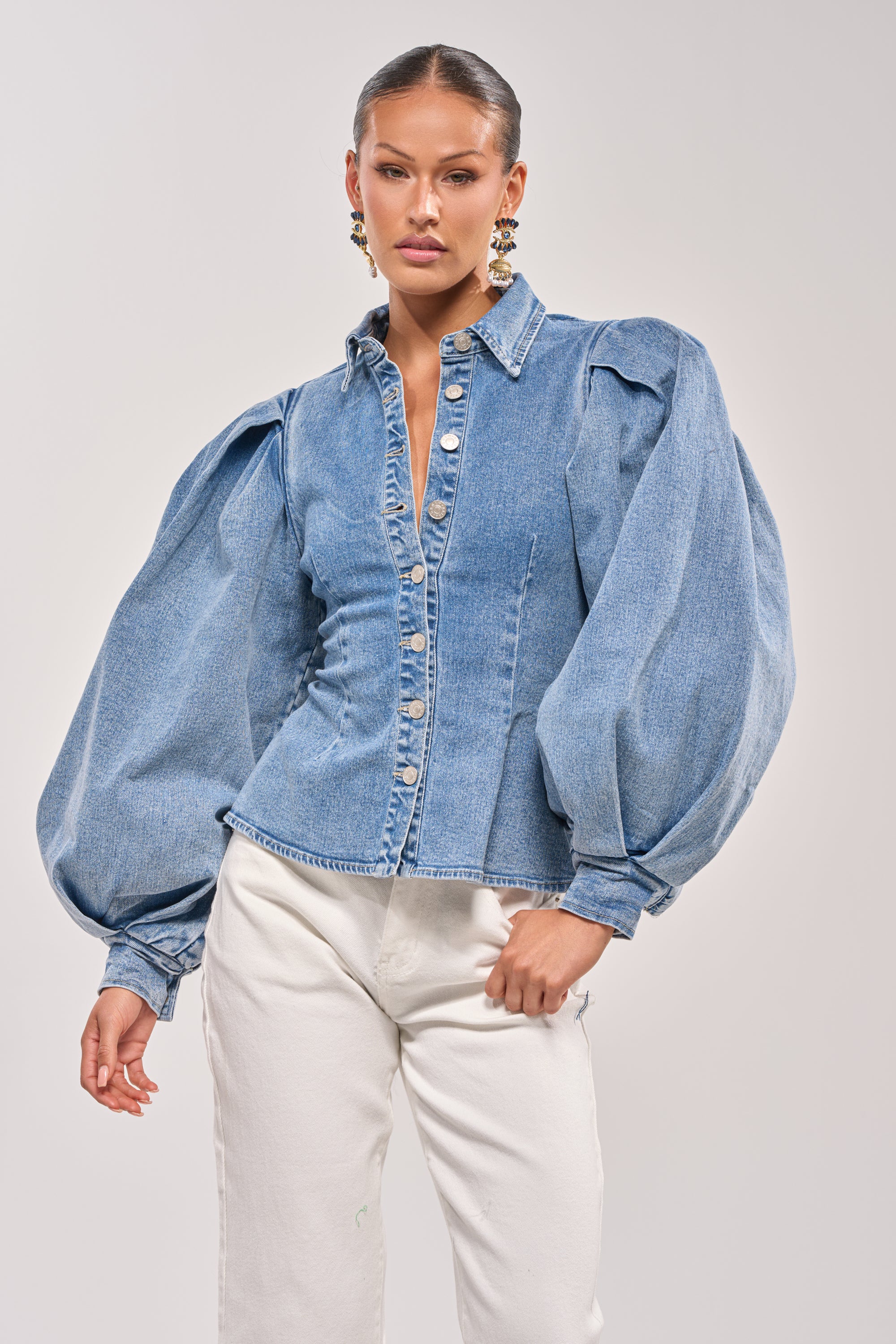 A woman stands confidently against a plain light background, wearing the JULIANA DENIM BLOUSE with voluminous puffed sleeves and white pants. 0