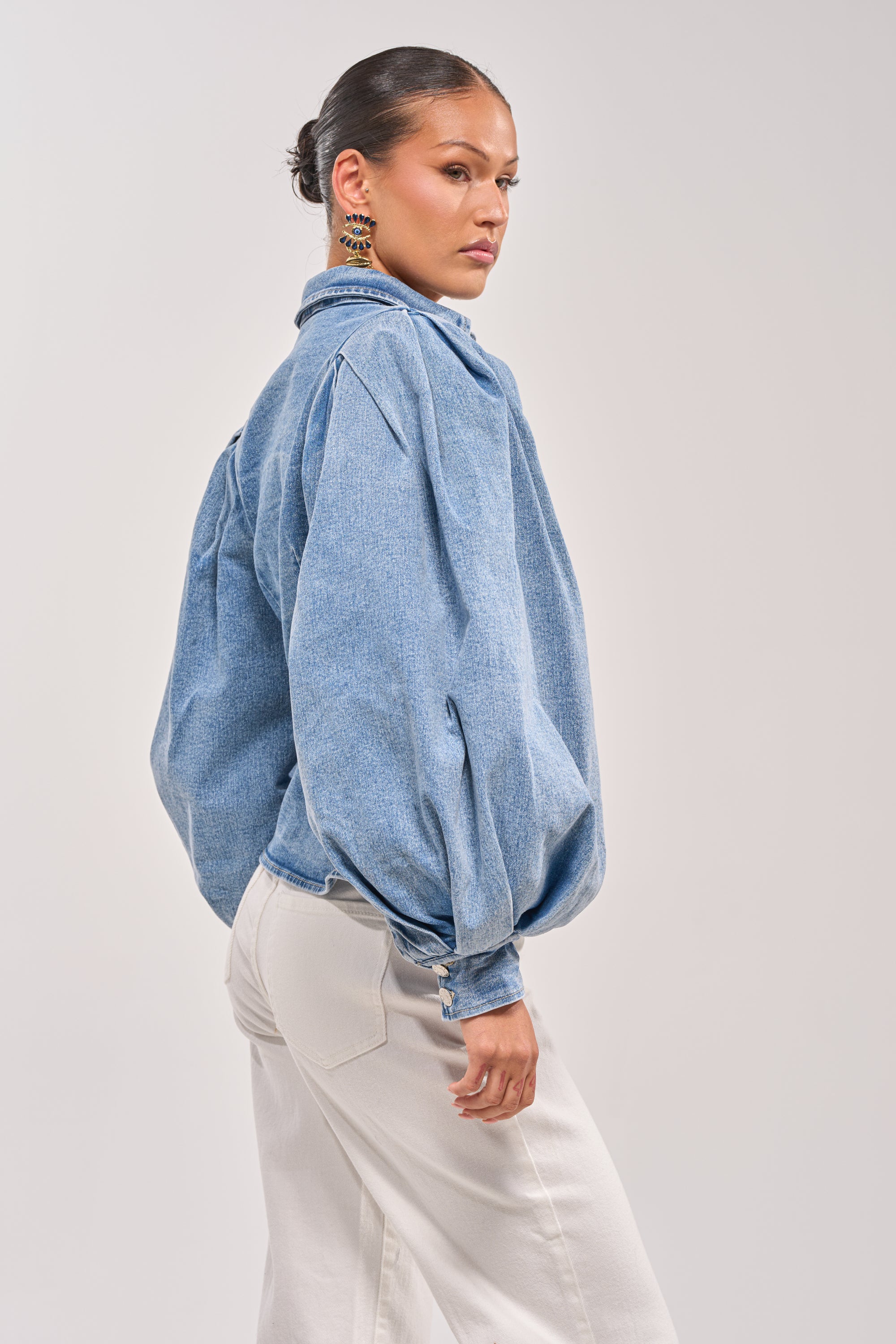 A woman with her hair in a low bun models the JULIANA DENIM BLOUSE, a loose-fitting, light blue button-down with puffed sleeves, paired with off-white pants. She stands sideways, looking over her shoulder against a plain background. 3