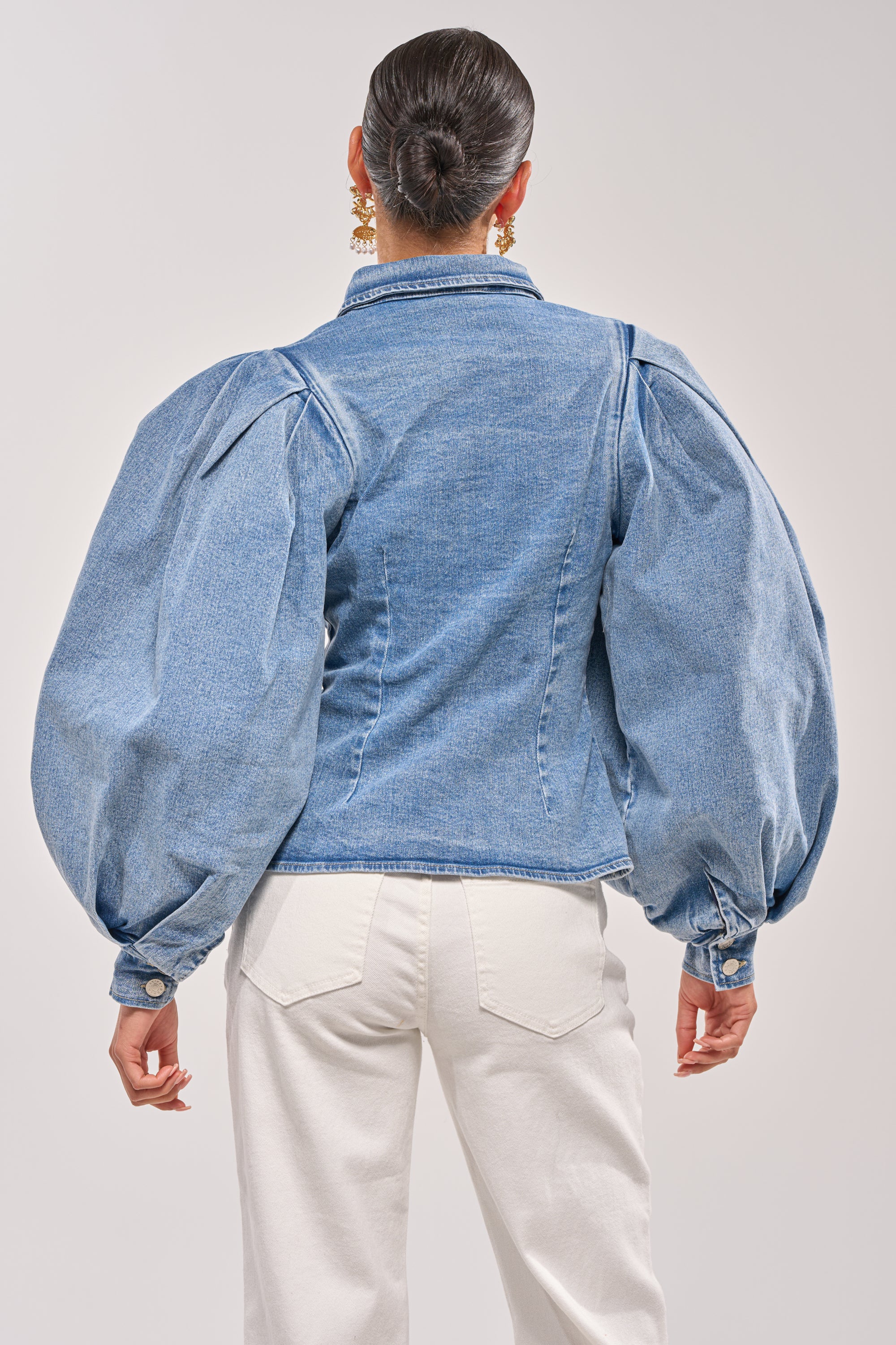 A person with dark hair in a sleek bun is seen from the back, wearing the JULIANA DENIM BLOUSE with dramatic puffed sleeves and white pants against a plain light background. 4