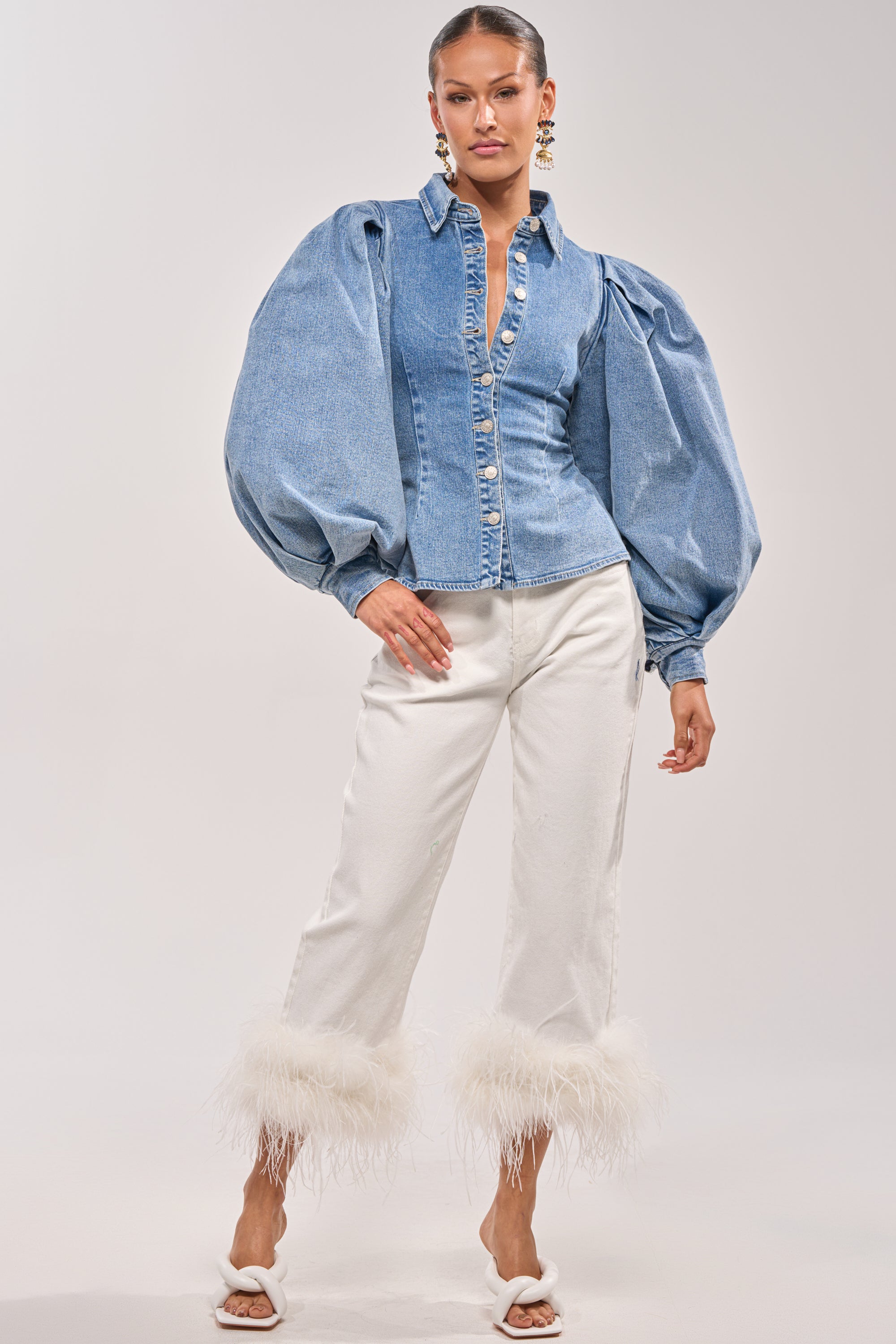 A woman poses in the JULIANA DENIM BLOUSE with balloon sleeves, white cropped feather-hem pants, white heeled sandals, and statement earrings against a plain background. 5