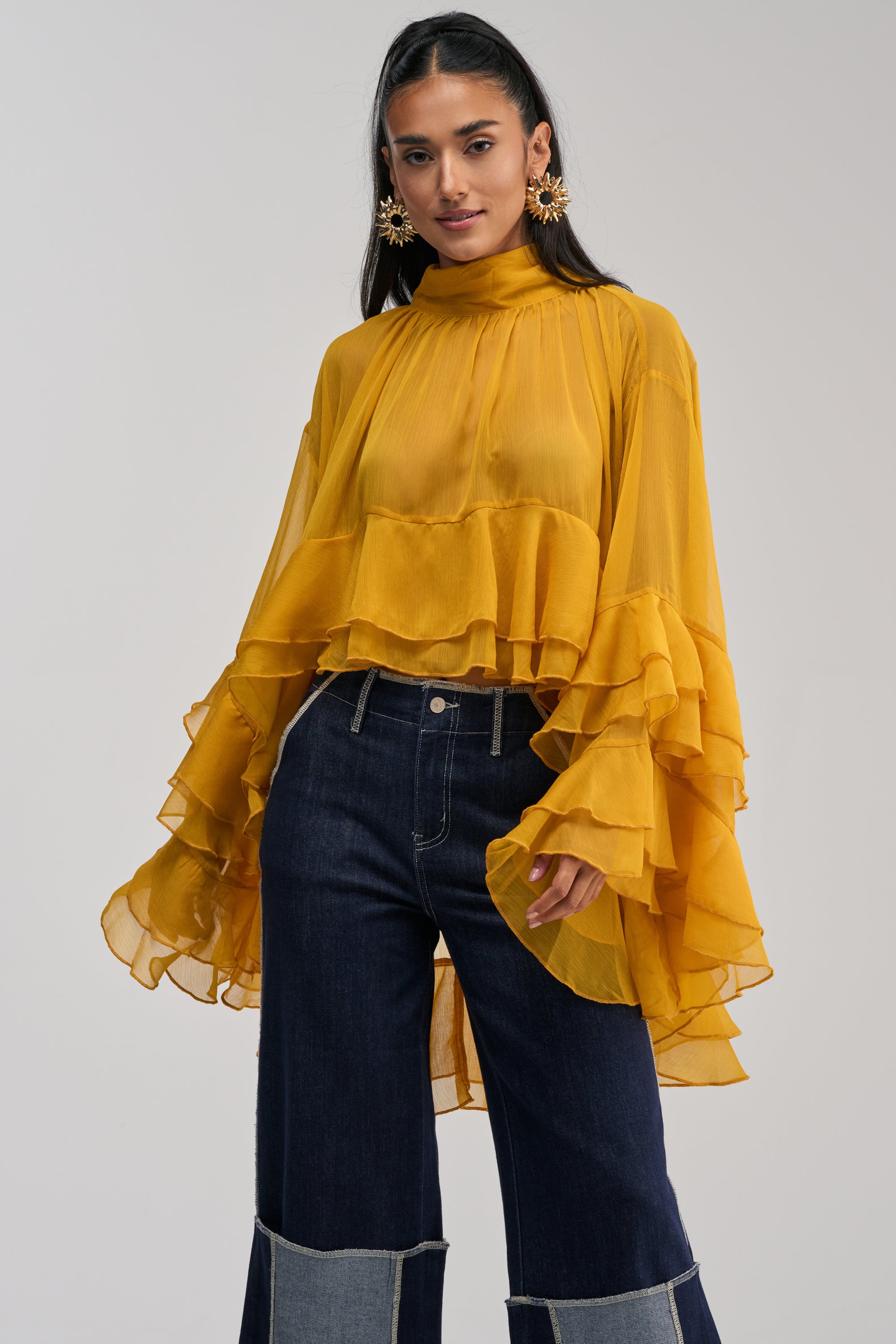 A woman wearing the MARGOT CHIFFON BLOUSE IN MUSTARD—an airy sheer blouse with tiered ruffles, dramatic flared sleeves, and a mock neckline—paired with dark wide-leg jeans featuring light denim panels, stands against a plain light background.