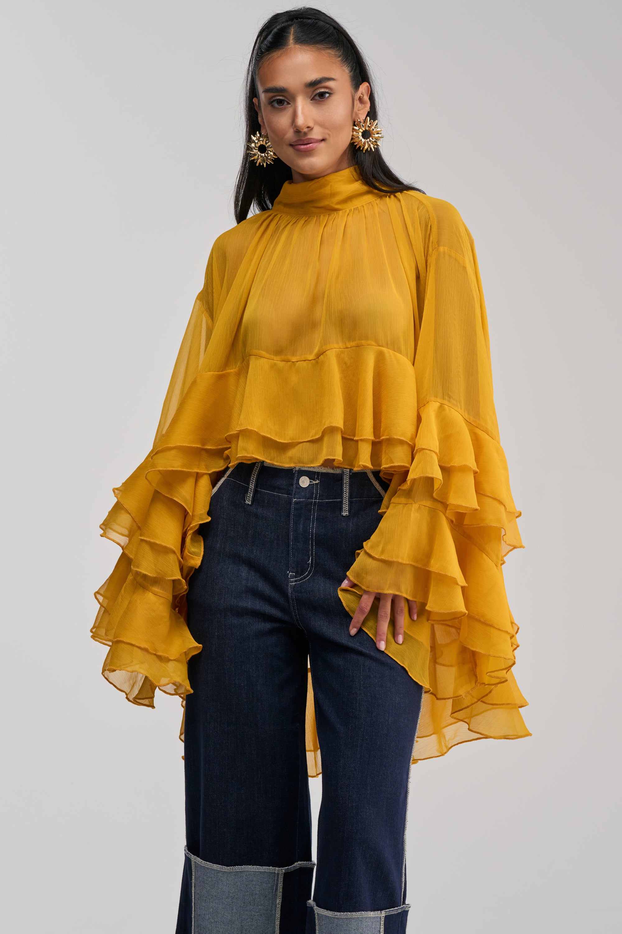A woman styled in the MARGOT CHIFFON BLOUSE IN MUSTARD, featuring tiered ruffles, dramatic ruffled sleeves, and a mock neckline, paired with dark wide-leg jeans and gold sunburst earrings.
