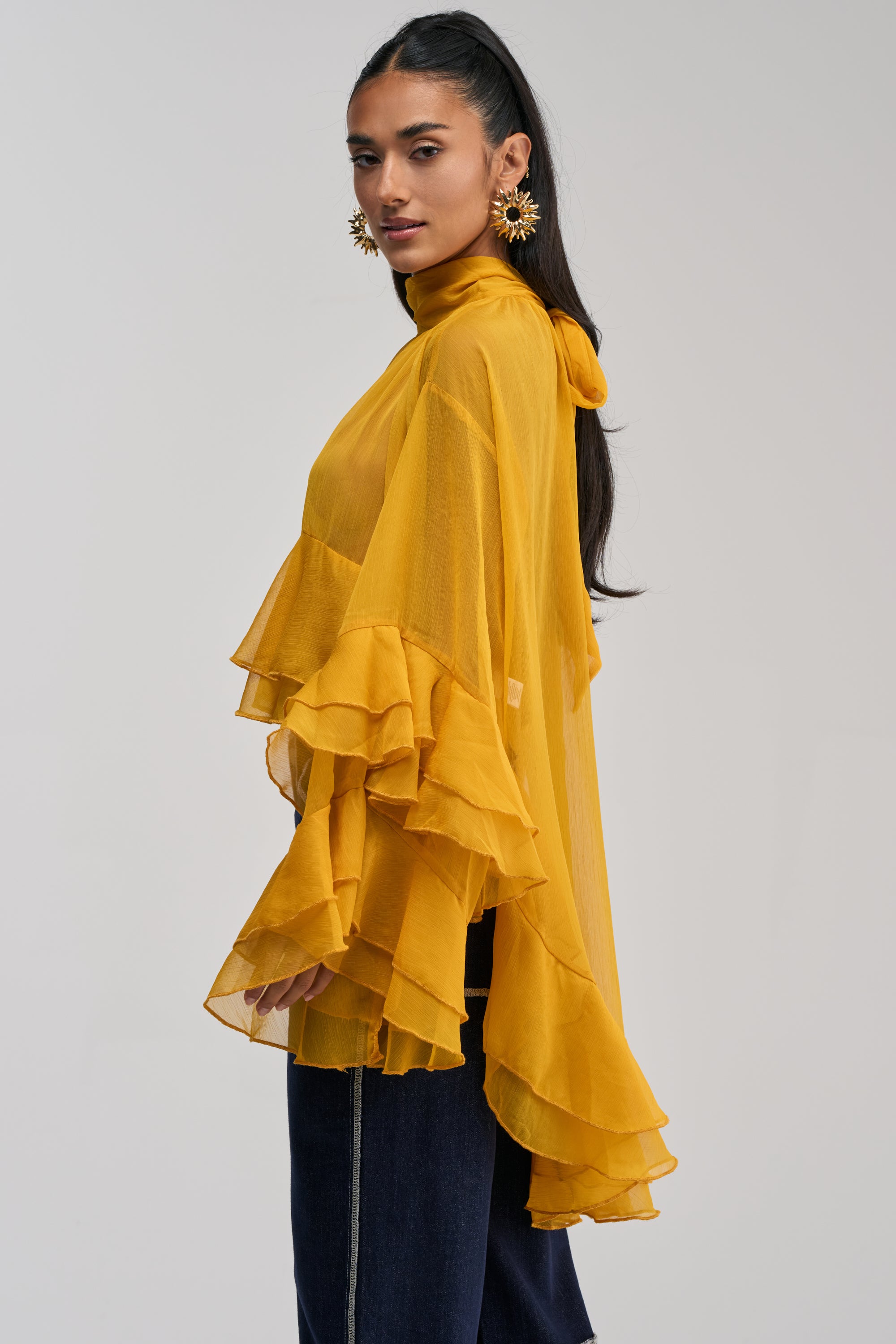 A woman stands sideways against a plain background, wearing the MARGOT CHIFFON BLOUSE IN MUSTARD—featuring flowy chiffon fabric, tiered ruffle sleeves, and a mock neckline—paired with statement earrings.