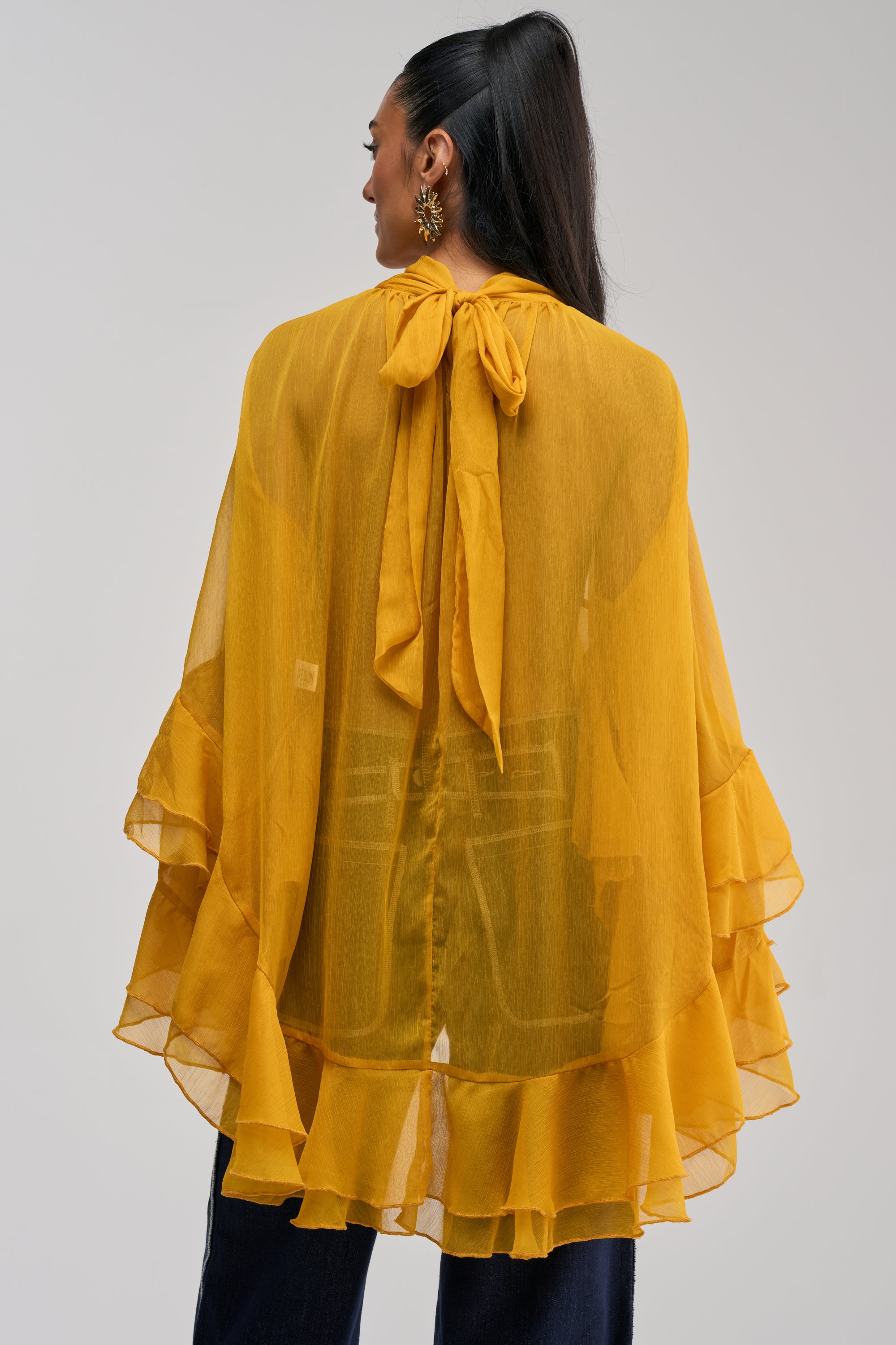 A woman is seen from behind wearing the MARGOT CHIFFON BLOUSE IN MUSTARD, featuring a sheer, ruffled cape detail and a tie neck, paired with dark navy trousers. The tiered ruffle design of the blouse complements her long dark hair styled in a high ponytail, along with gold earrings.