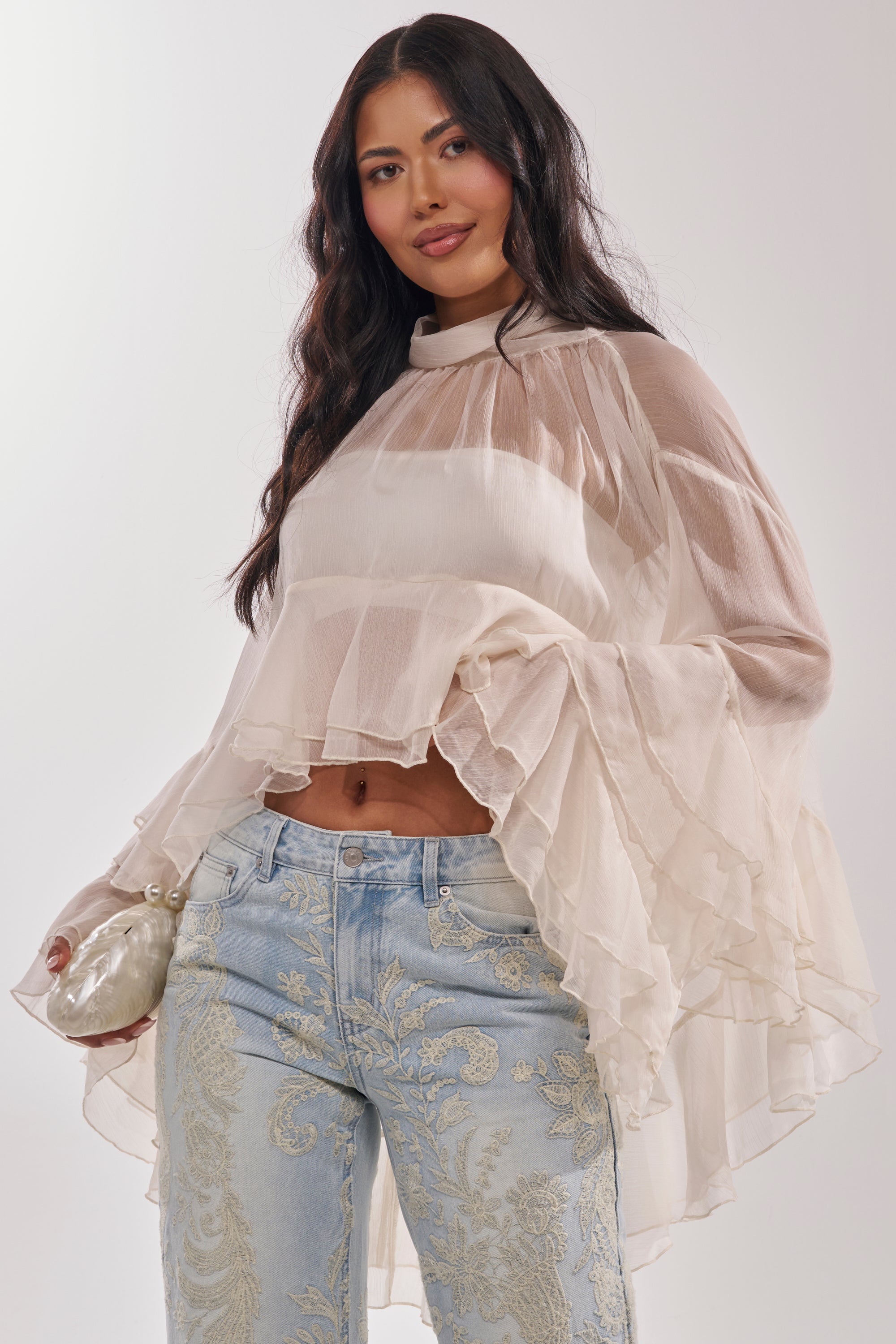 A woman with long dark hair wears the MARGOT CHIFFON BLOUSE—sheer with tiered ruffled sleeves—paired with high-waisted light blue embroidered jeans and holds a small pearlescent clutch. 2