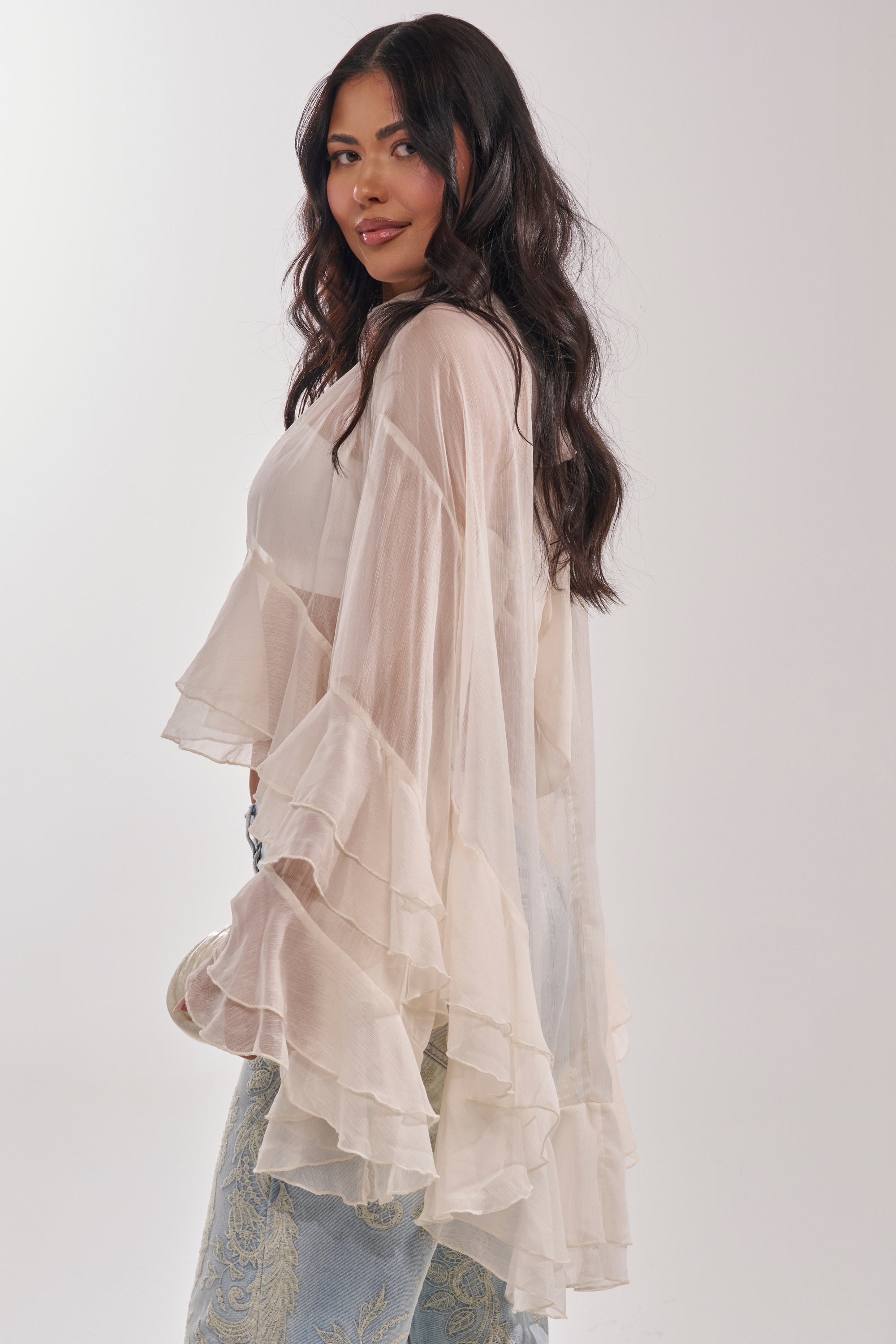 A woman with long, wavy dark hair models the MARGOT CHIFFON BLOUSE, featuring flowing tiered ruffles, paired with light blue patterned pants. She stands sideways and looks over her shoulder against a plain background. 3