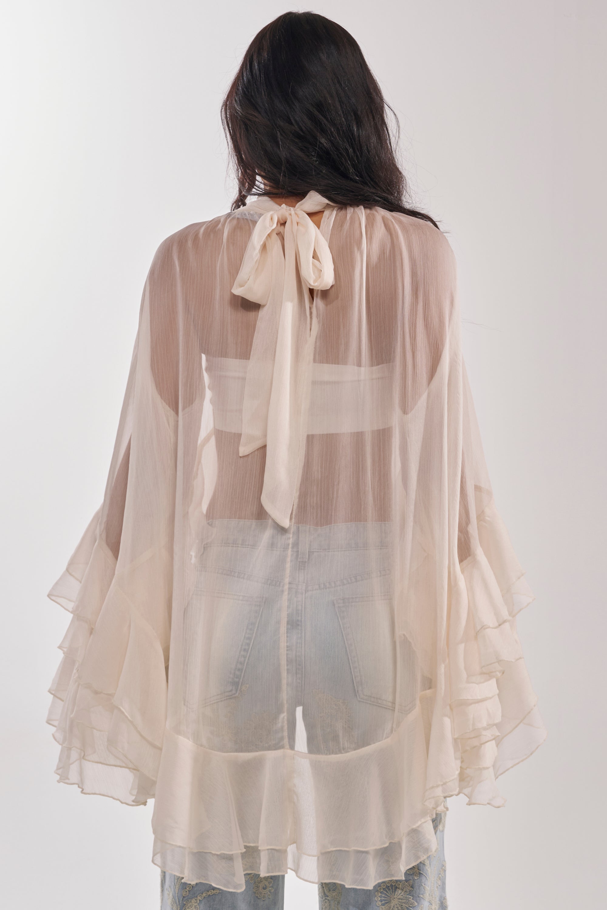 A woman faces away, wearing the MARGOT CHIFFON BLOUSE—sheer cream fabric with tiered ruffle sleeves and a bow at the mock neckline. Denim jeans are visible beneath the translucent blouse. 4