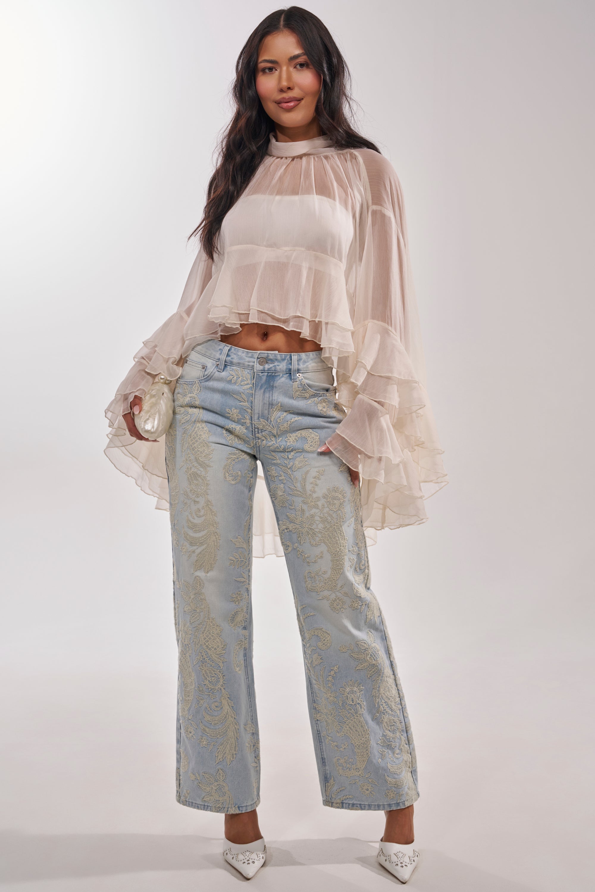 A woman with long dark hair slightly smiling wears the MARGOT CHIFFON BLOUSE, sheer with tiered ruffles, paired with light blue paisley jeans, white pointed heels, and holds a small white clutch. 5