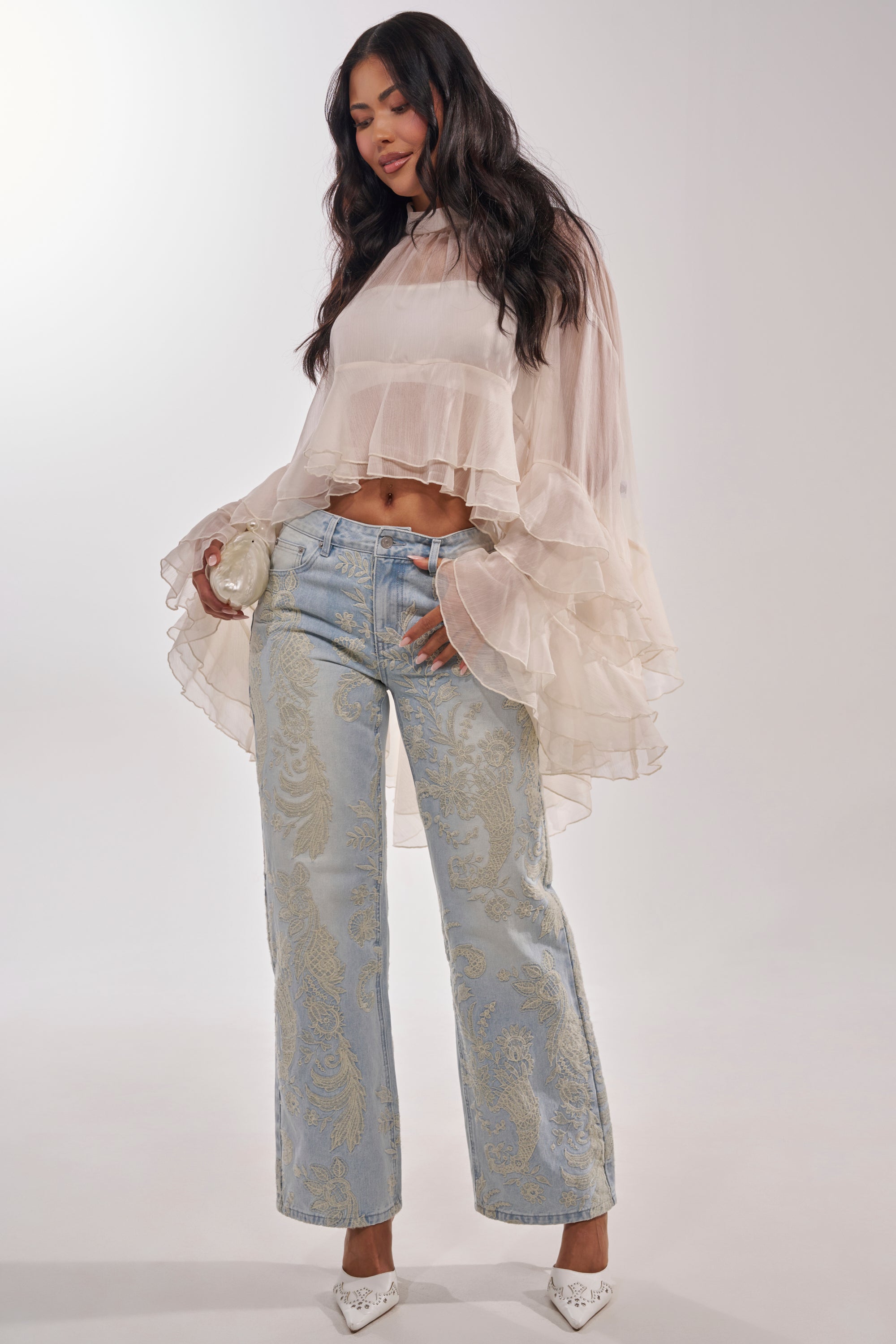A woman models the MARGOT CHIFFON BLOUSE with tiered ruffles and flared sleeves, paired with light blue jeans featuring gold paisley designs and white pointed-toe heels, holding a round purse against a plain white background. 6