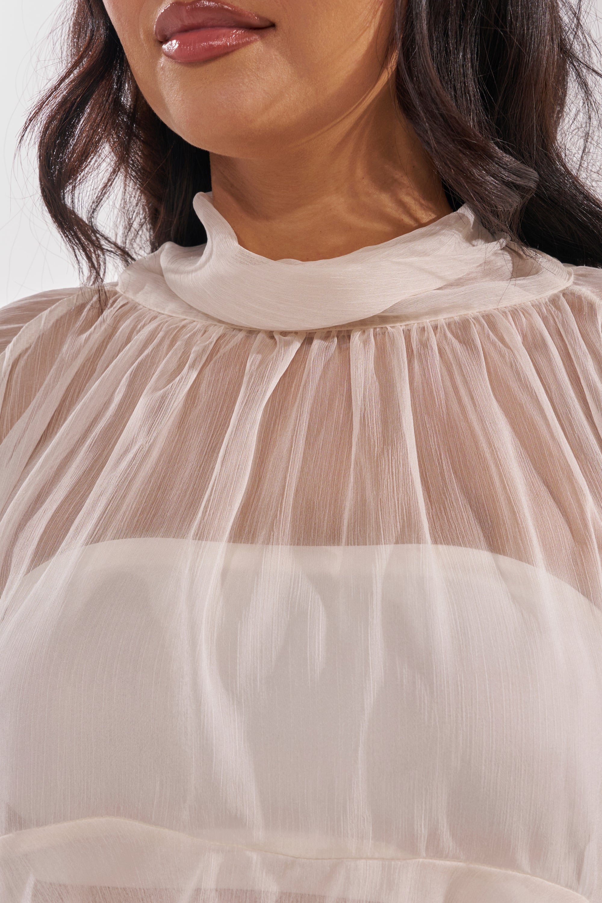 A close-up of a woman wearing the MARGOT CHIFFON BLOUSE, featuring a sheer finish, high mock neckline, and gathered details. Only her lower face and upper torso are visible. 8