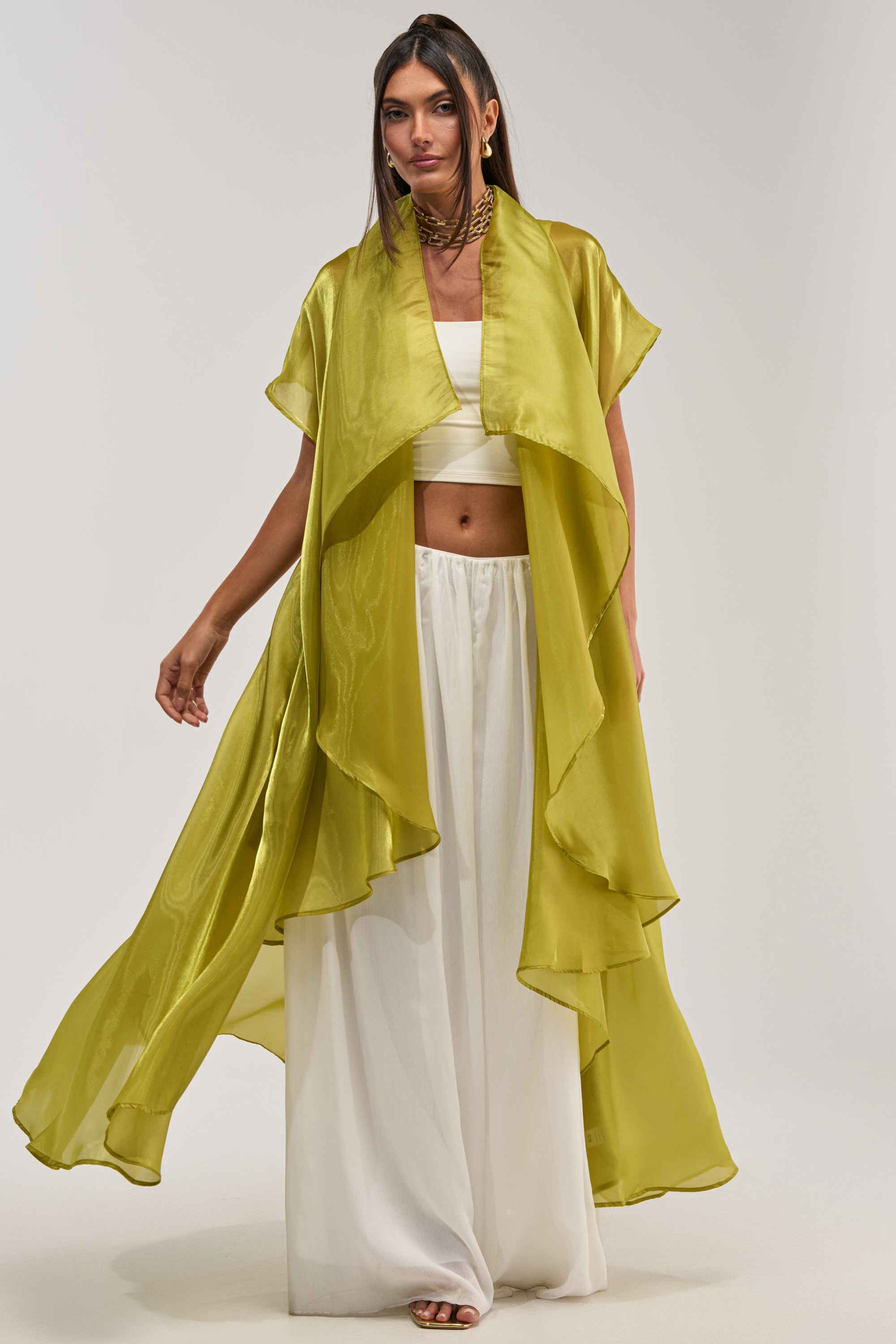 A woman wears the JULIETTE CHIFFON BLOUSE, a sheer, flowing chartreuse duster over a white crop top and wide-leg pants. Posed against a light background, her maxi-length look is elegant and modern.