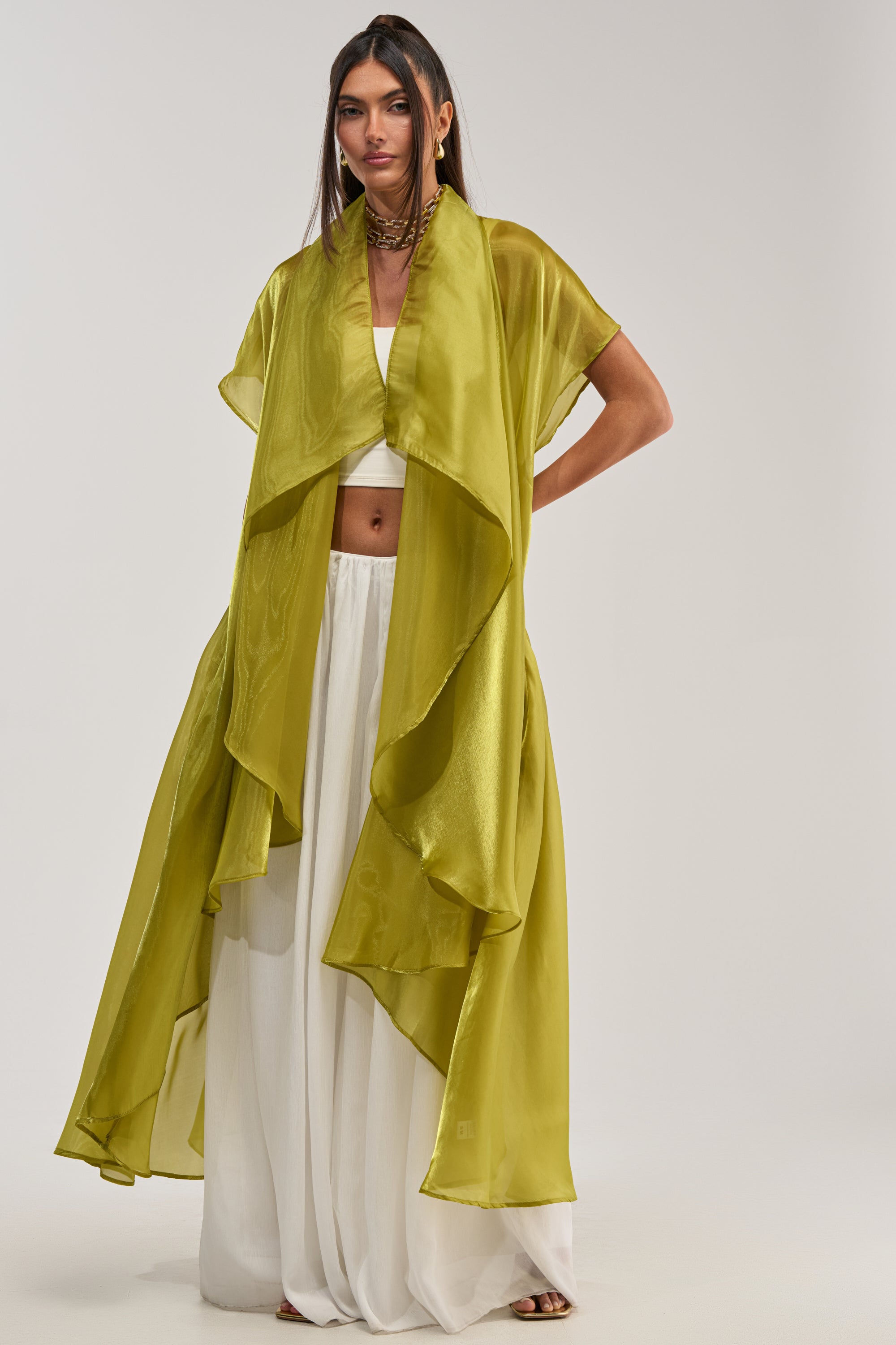 A woman stands against a plain light background in the flowy, sheer lime green JULIETTE CHIFFON BLOUSE styled open over a white crop top and wide-legged white pants—a chic outfit perfect for special occasions.