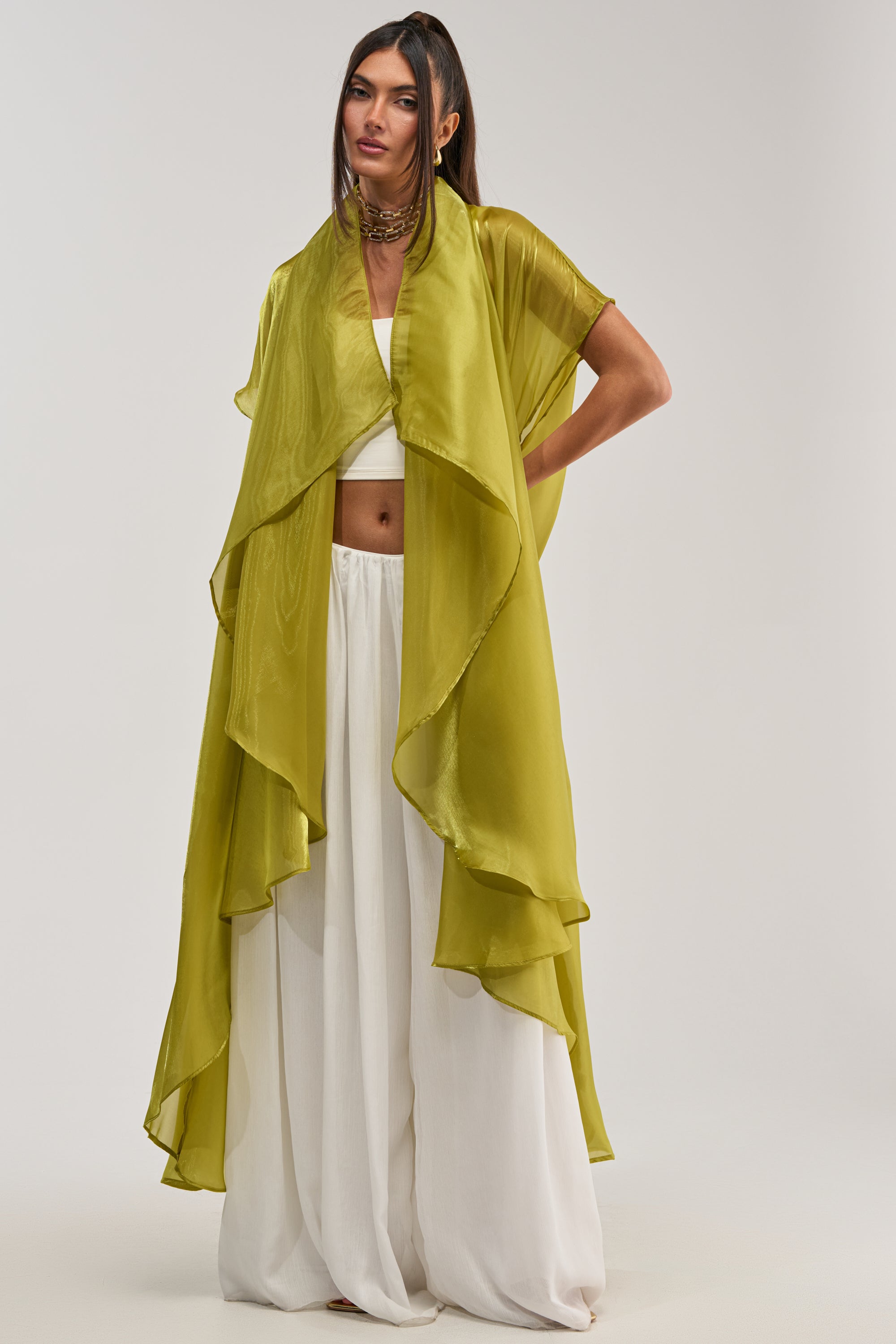 A woman models the JULIETTE CHIFFON BLOUSE—a sheer, layered olive green piece—styled over a white crop top and wide-legged pants against a light background, offering chic inspiration for occasion dressing.