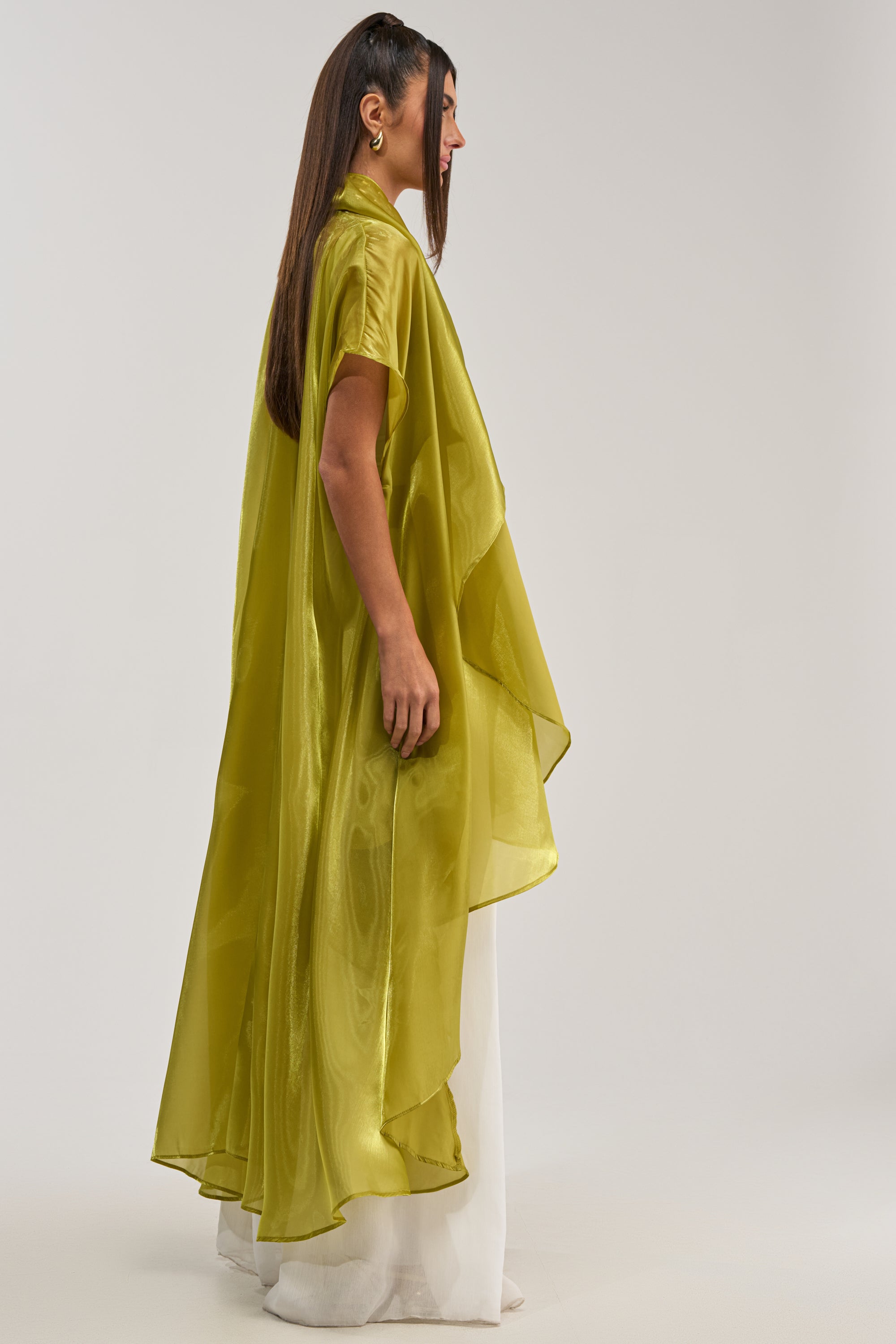 A woman in profile stands against a plain background, wearing the JULIETTE CHIFFON BLOUSE in flowy lime green chiffon over a white outfit. Her straight hair is tucked behind one ear, with her hands resting at her sides.