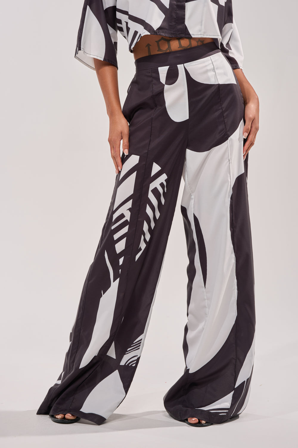 A person stands barefoot with hands on thighs, wearing the NOIR GODDESS PALAZZO PANT and a matching top featuring bold black and white abstract designs—ideal for relaxed vacation wear against a plain, light-colored background. 0