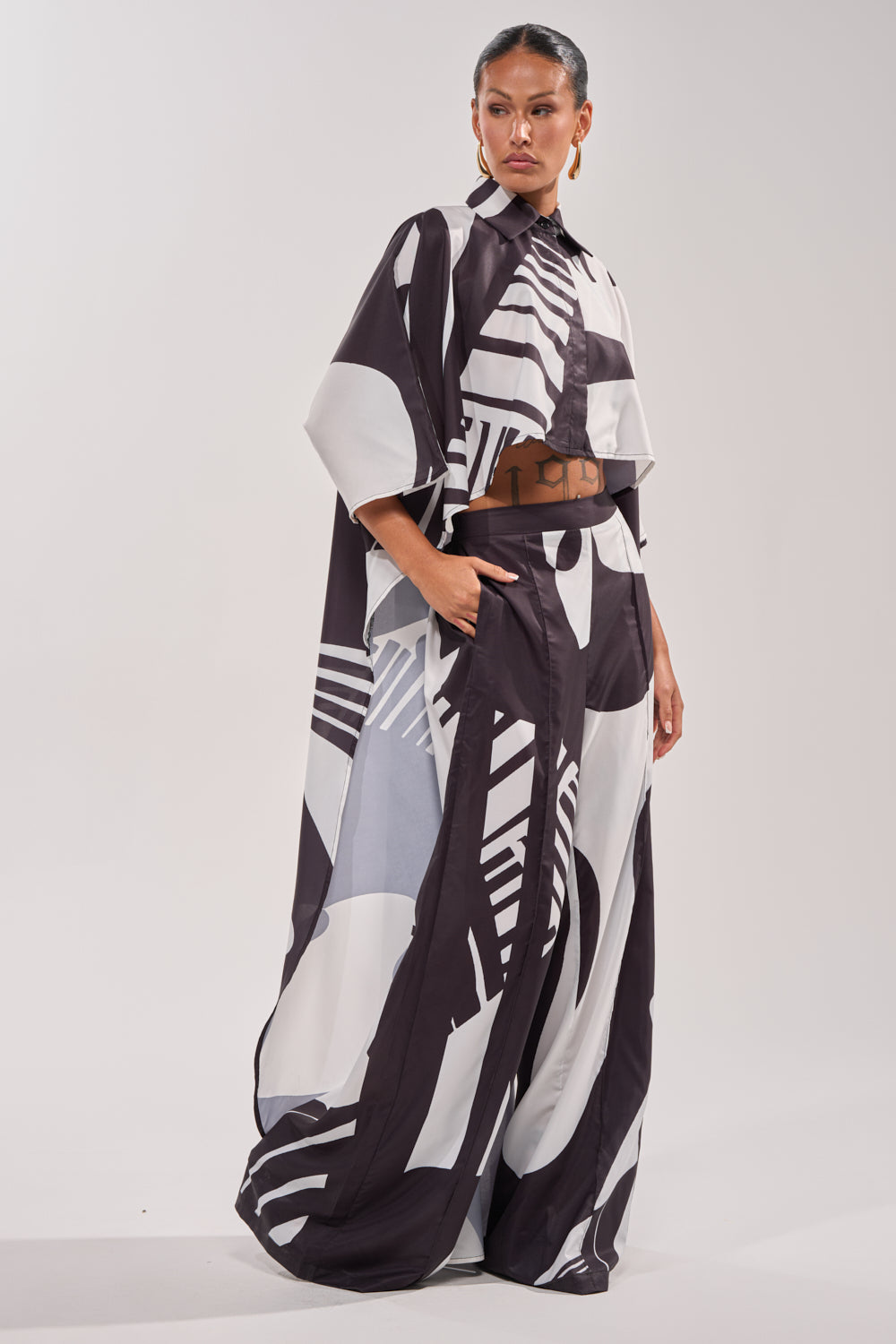 A woman stands confidently in the NOIR GODDESS PALAZZO PANT, a loose, black and white geometric-patterned outfit with wide sleeves and pockets, hair pulled back against a light background—chic vacation style. 8