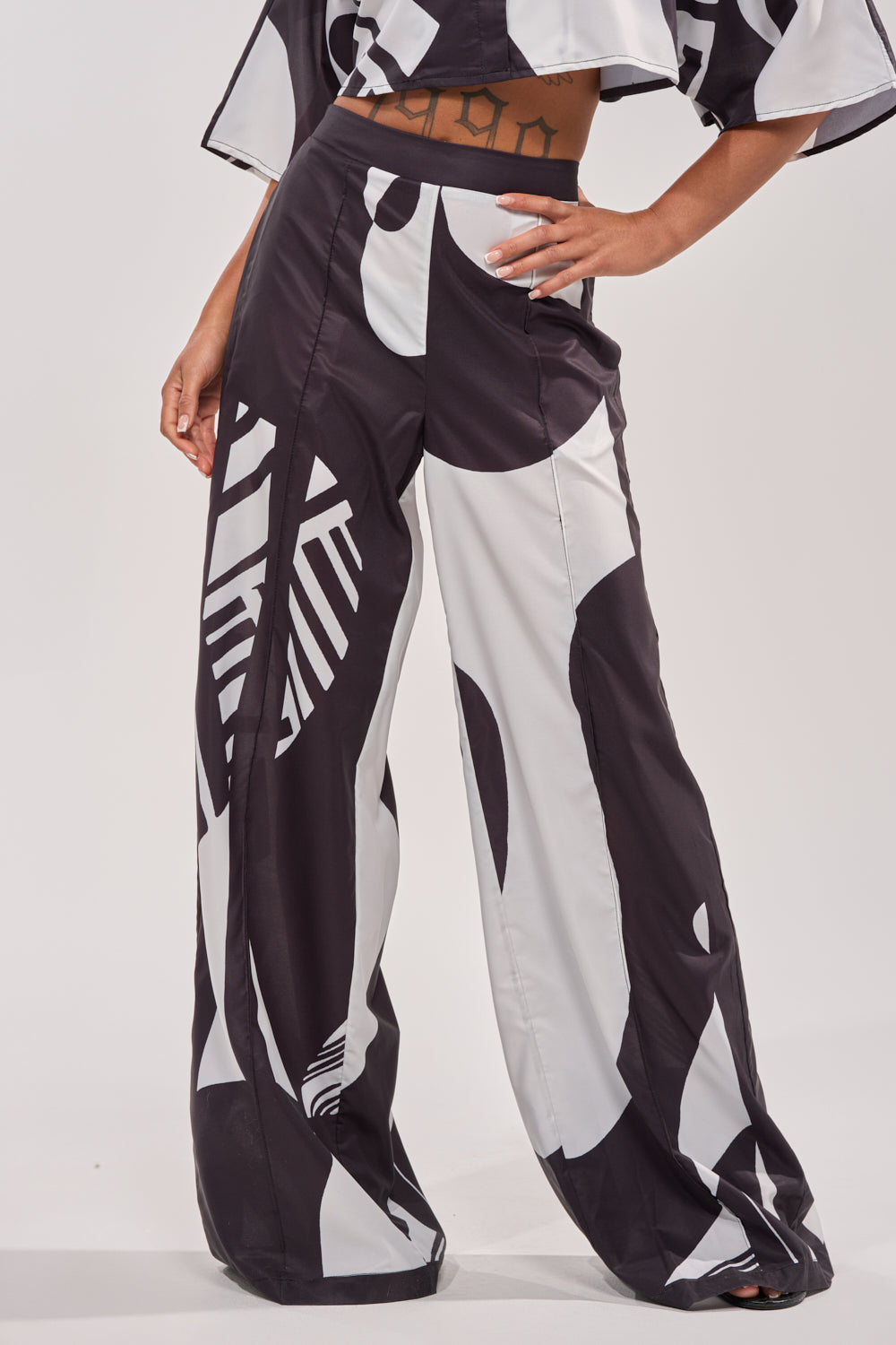 A person poses with one hand on their hip, wearing the NOIR GODDESS PALAZZO PANT—high-waisted black and white pants with bold abstract patterns, ideal for vacation style. Their midriff and tattoo are partially visible. 1