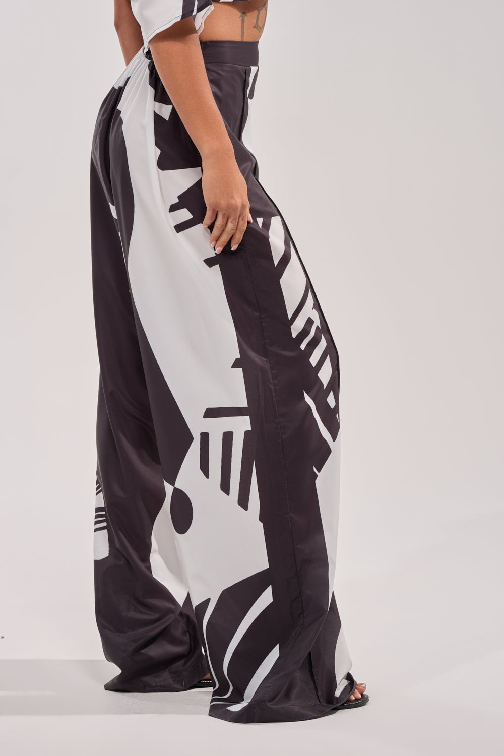 Only the lower body of a person in NOIR GODDESS PALAZZO PANT—loose palazzo pants featuring a bold black and white graphic print—is visible as they stand sideways against a plain white background. 2