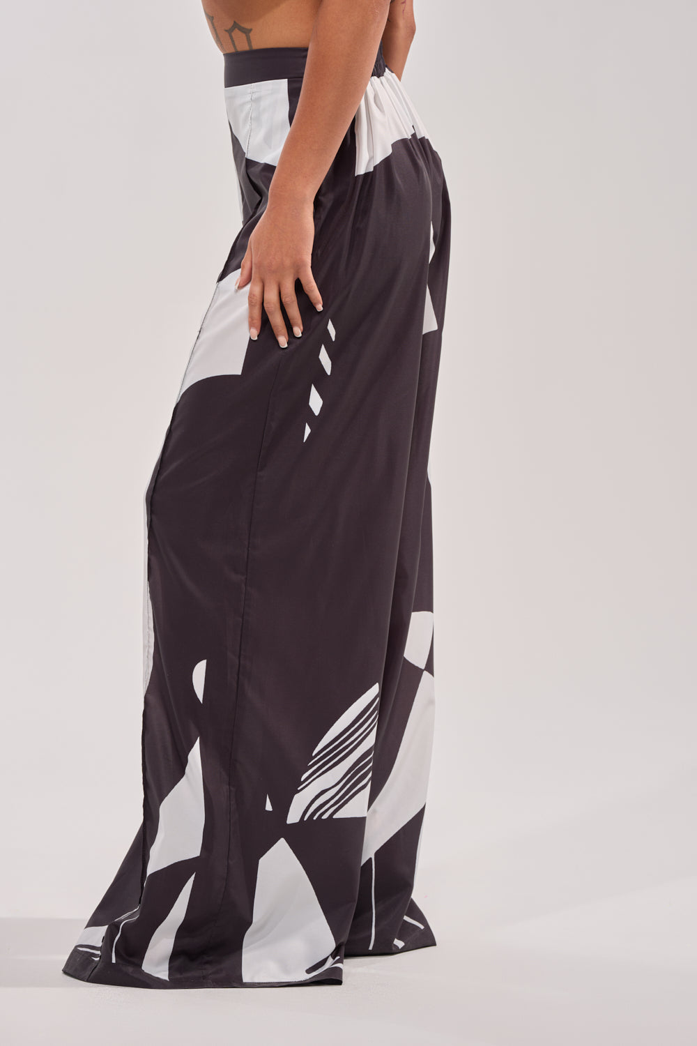 A person stands sideways in the NOIR GODDESS PALAZZO PANT, featuring bold black and white abstract patterns. The image is cropped from mid-torso down, highlighting the wide-leg silhouette against a plain light gray background. 3