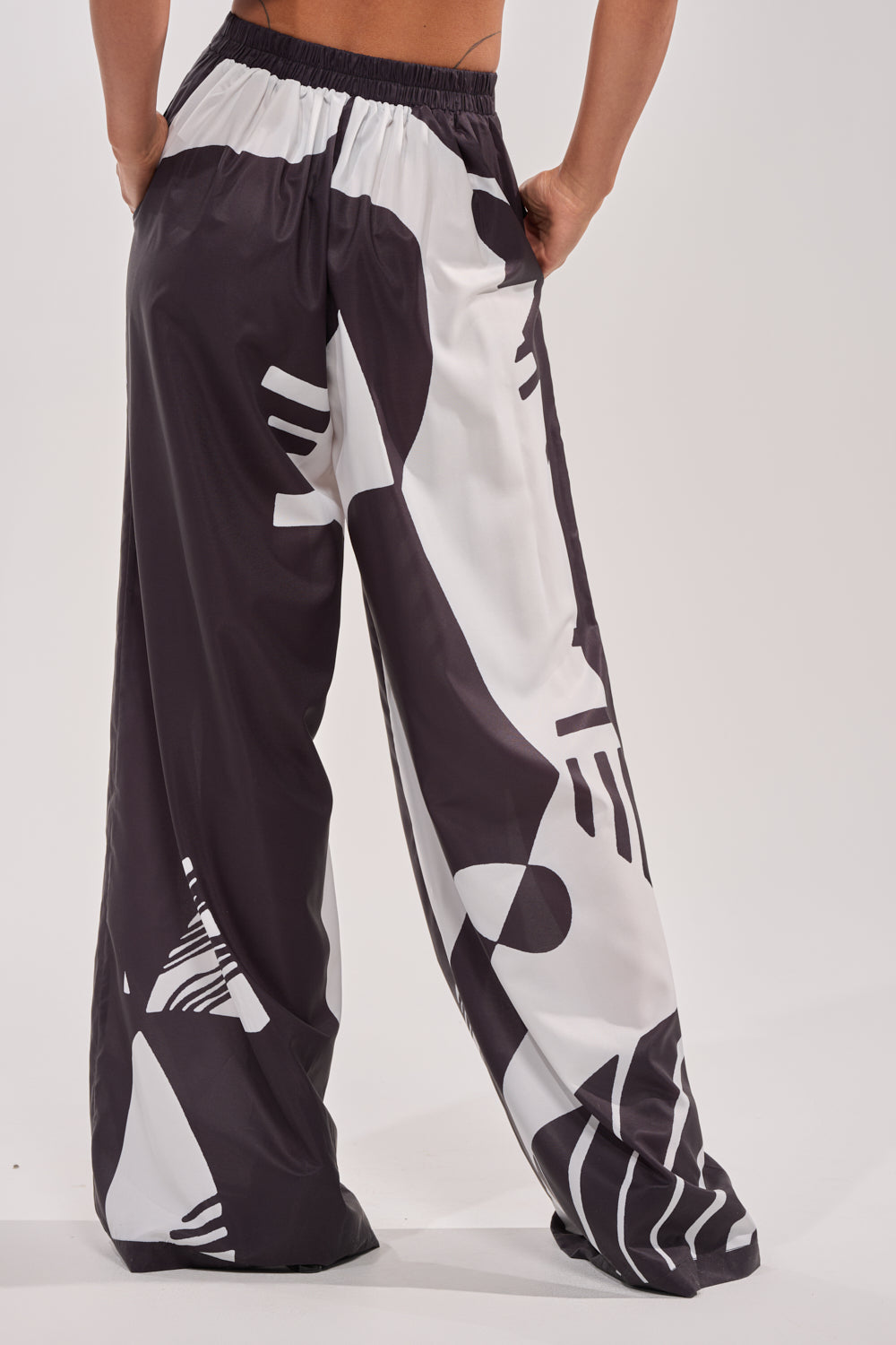 A person stands with hands in pockets, wearing NOIR GODDESS PALAZZO PANT—loose black and white palazzo pants featuring an abstract pattern, relaxed fit, and elastic waistband. Photo captured from behind highlights their perfect vacation style. 4