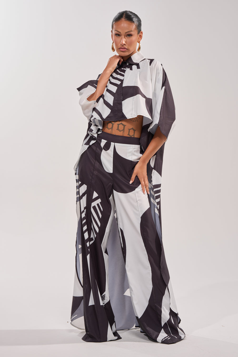 A woman stands confidently in NOIR GODDESS PALAZZO PANT paired with a flowing geometric top and large hoop earrings—an effortlessly chic vacation look on a plain backdrop. 5