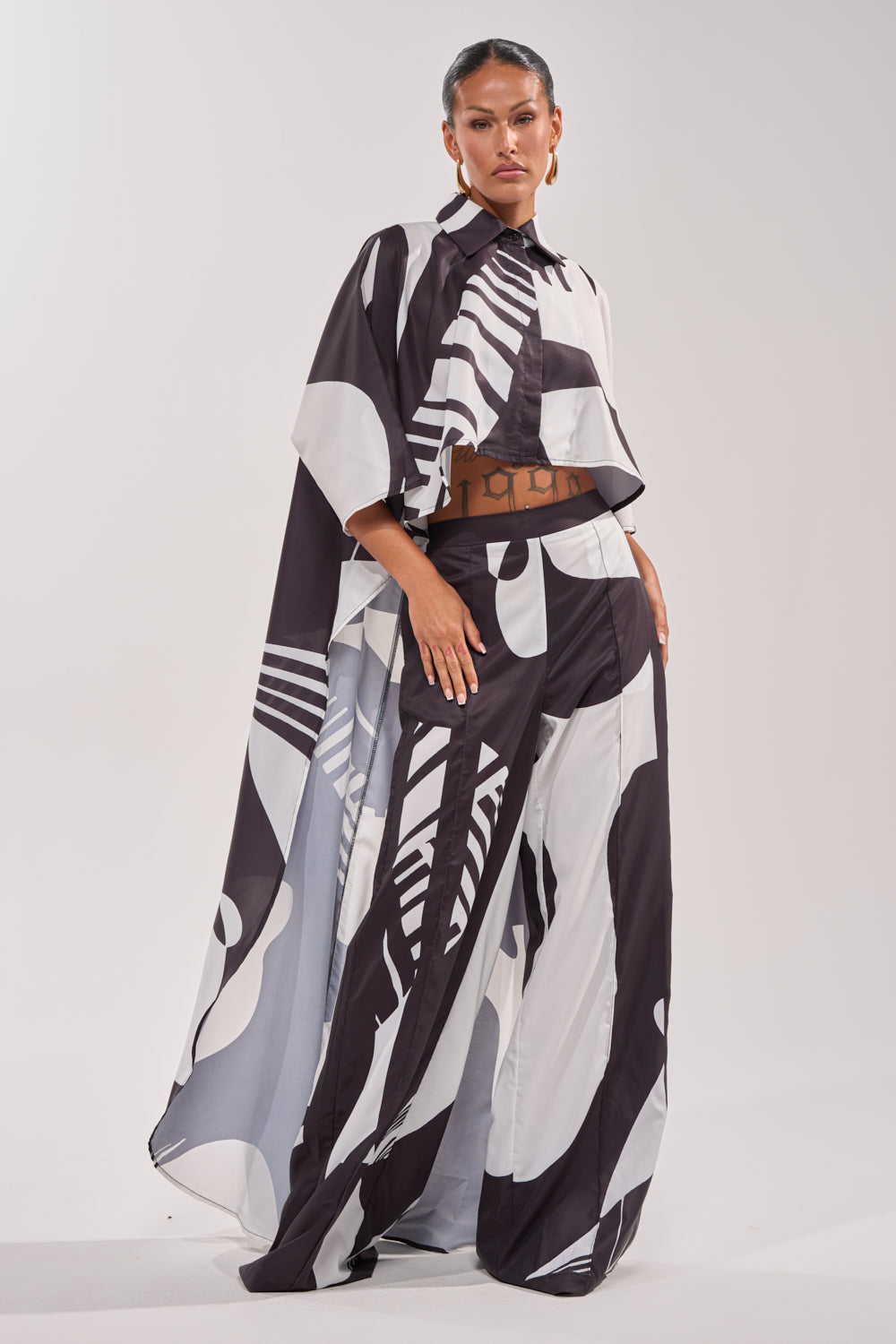 A woman confidently poses in NOIR GODDESS PALAZZO PANT and a cape-style top—an abstract, vacation-ready look—set against a simple light background. 6