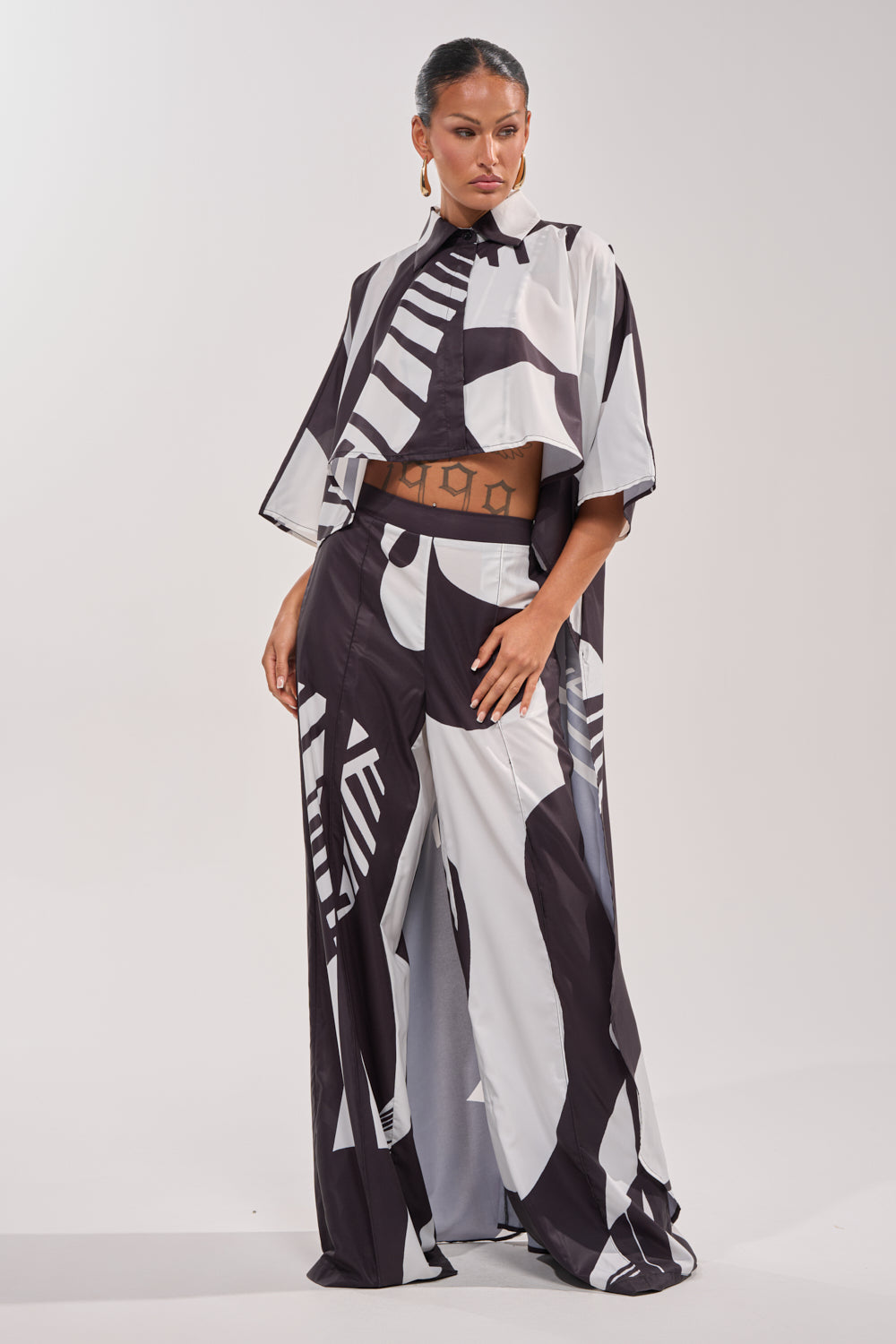 A woman exudes confidence in vacation wear—a NOIR GODDESS PALAZZO PANT set with a loose, black-and-white design and a cropped, short-sleeve button-up shirt that reveals her midriff and geometric stomach tattoos. 7