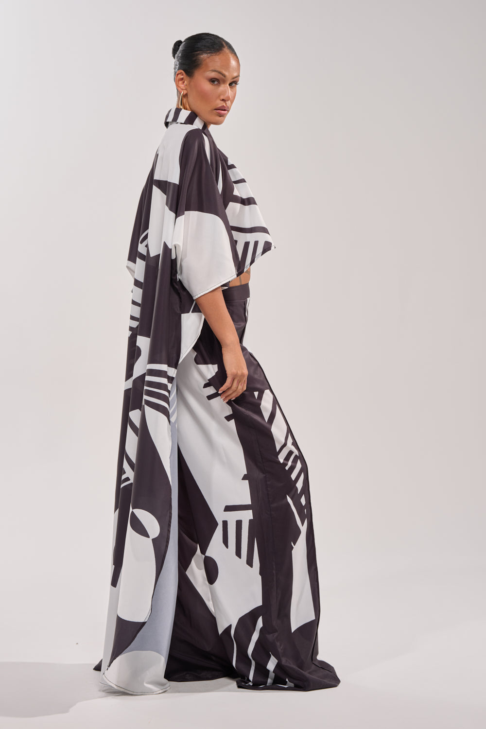 A woman stands in profile wearing the NOIR GODDESS COLLARED CAPE TOP—an elegant, long resort wear piece with bold black and white geometric patterns. Her hair is styled in a sleek bun as she poses confidently against a plain background. 4