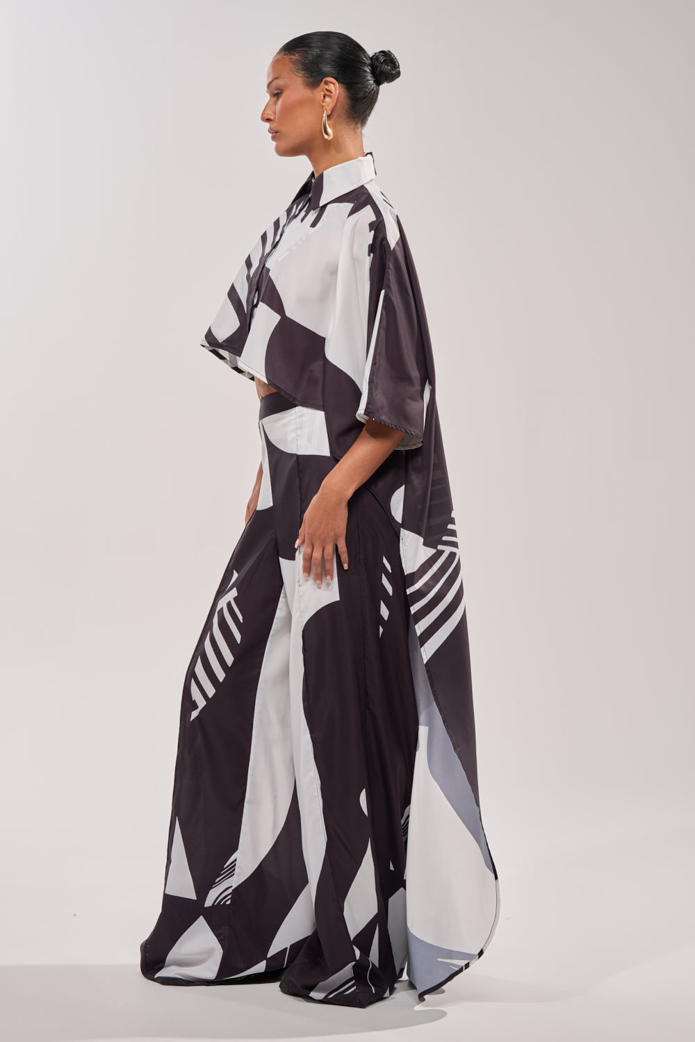 A woman stands in profile wearing the NOIR GODDESS COLLARED CAPE TOP, a loose black and white print blouse styled as resort wear with a long skirt. Her hair is in a sleek bun, accented by large hoop earrings against a light background. 5