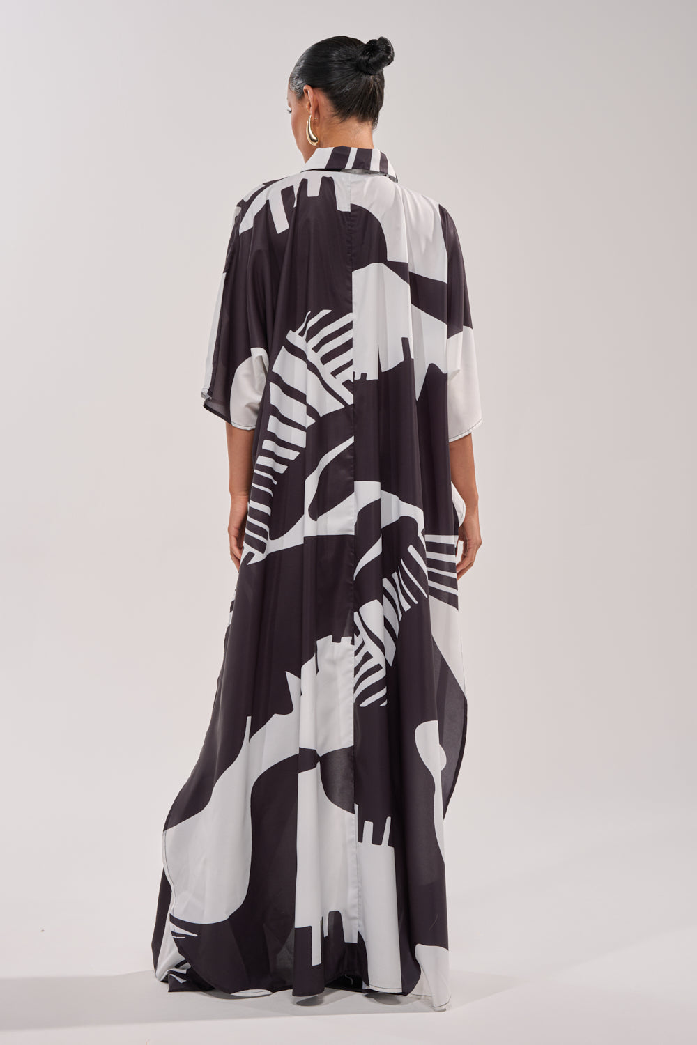 A person with dark hair in a bun faces away from the camera, wearing the NOIR GODDESS COLLARED CAPE TOP—a flowing, black and white patterned resort wear piece with short sleeves—against a plain light background. 6
