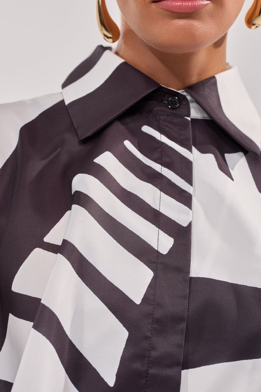 Close-up of a person wearing the NOIR GODDESS COLLARED CAPE TOP, featuring bold black and white abstract stripes—ideal for resort wear. The image shows only the lower face, neck, and part of a gold earring. 7