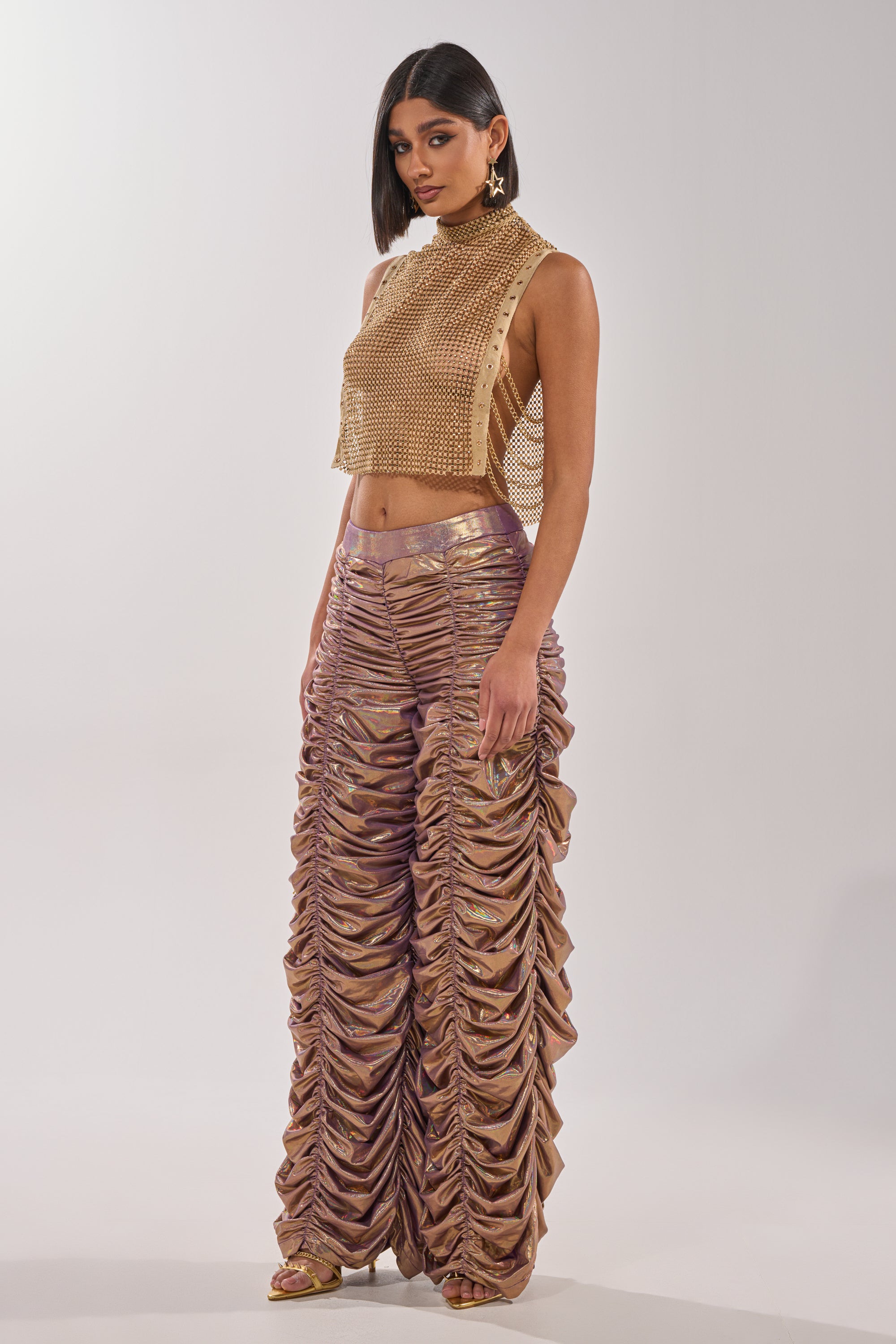 A woman wears the TEARS IN THE CLUB OPEN SIDES MOCK NECK RHINESTONE MESH BLOUSE—a sleeveless gold mesh top with side button details—paired with high-waisted shiny ruched gold and purple pants, statement earrings, gold heels, and straight dark hair. 9