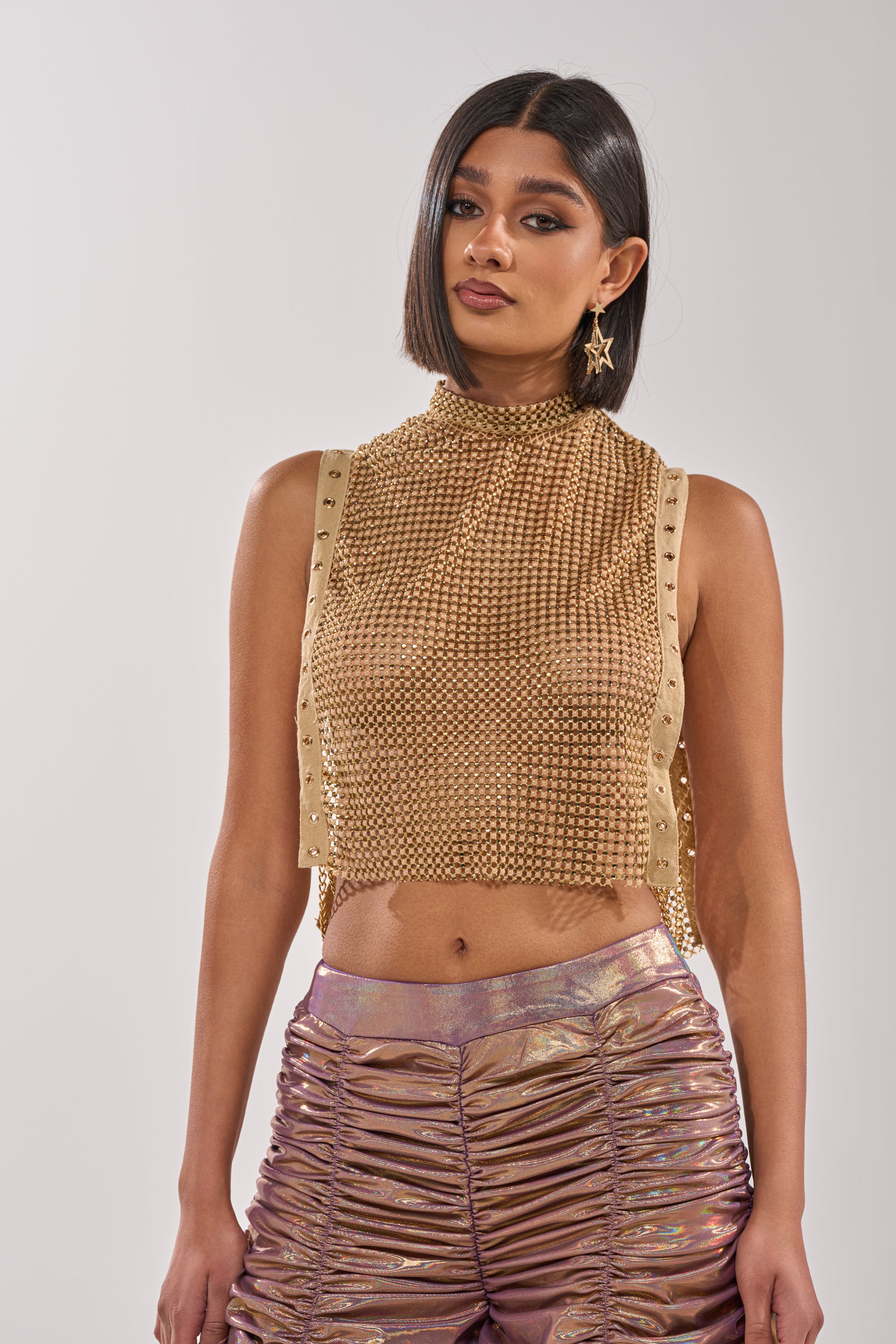 A woman with a sleek bob wears the TEARS IN THE CLUB OPEN SIDES MOCK NECK RHINESTONE MESH BLOUSE and high-waisted mauve ruched pants, accessorized with star-shaped chain link earrings, posing against a plain light background. 1