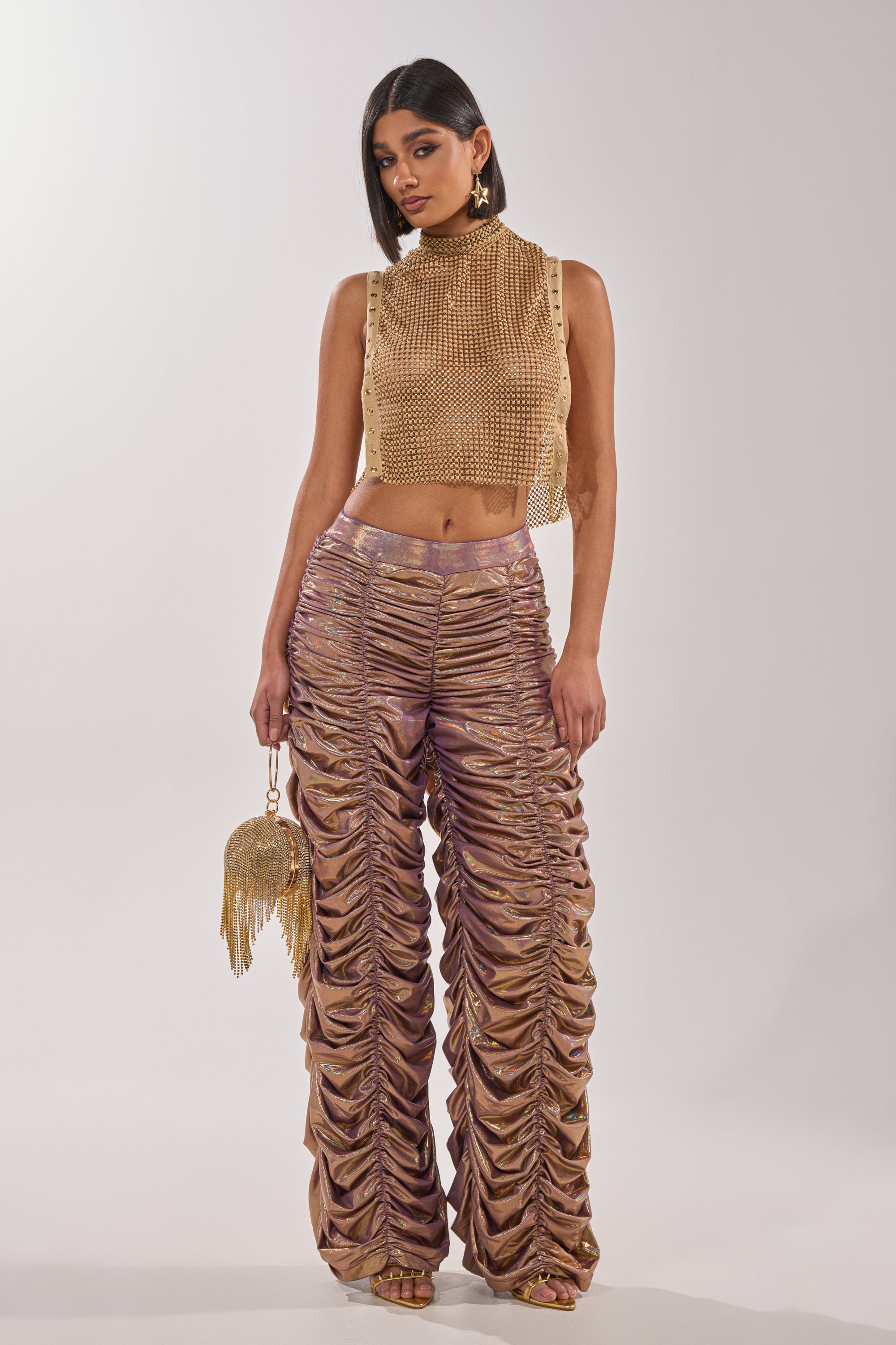 A woman poses in the TEARS IN THE CLUB OPEN SIDES MOCK NECK RHINESTONE MESH BLOUSE paired with shiny bronze ruched pants, a round gold tassel purse, gold earrings, and heels against a plain light background. 6
