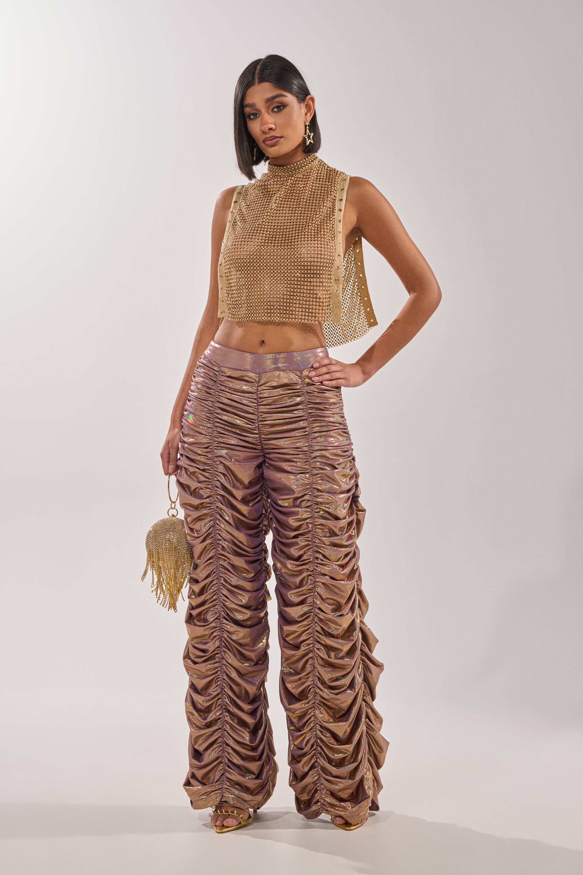 A woman confidently poses in the TEARS IN THE CLUB OPEN SIDES MOCK NECK RHINESTONE MESH BLOUSE, styled with metallic ruched wide-leg pants, a gold fringe purse, and statement earrings against a plain white background. 7