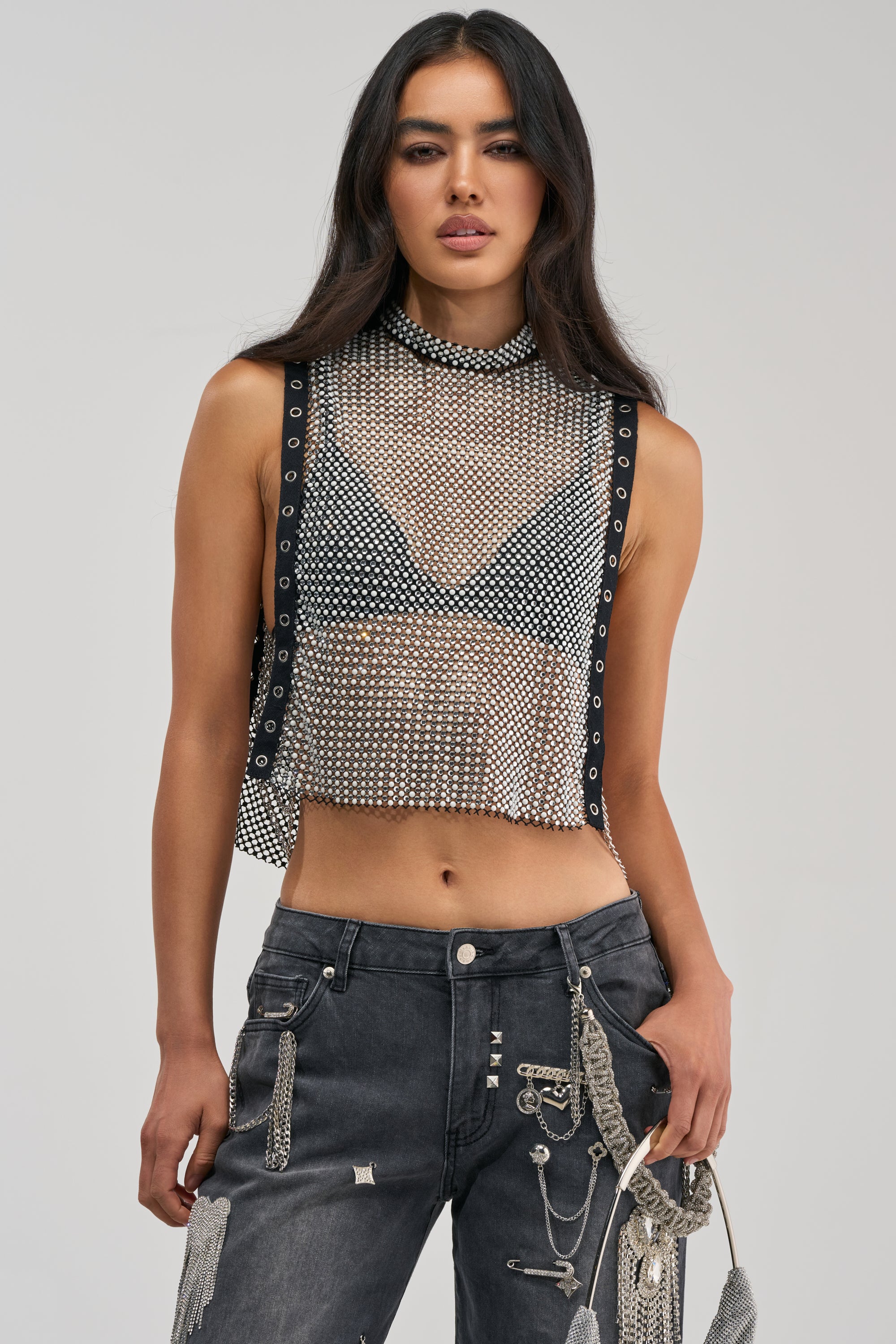 A woman with long dark hair stands against a plain light background, wearing the NO CRYING IN THE CLUB RHINESTONE MESH TOP—a sleeveless crop mesh statement piece layered over a black bra and paired with dark jeans adorned with silver chains and pins, perfect for a striking night-out look.
