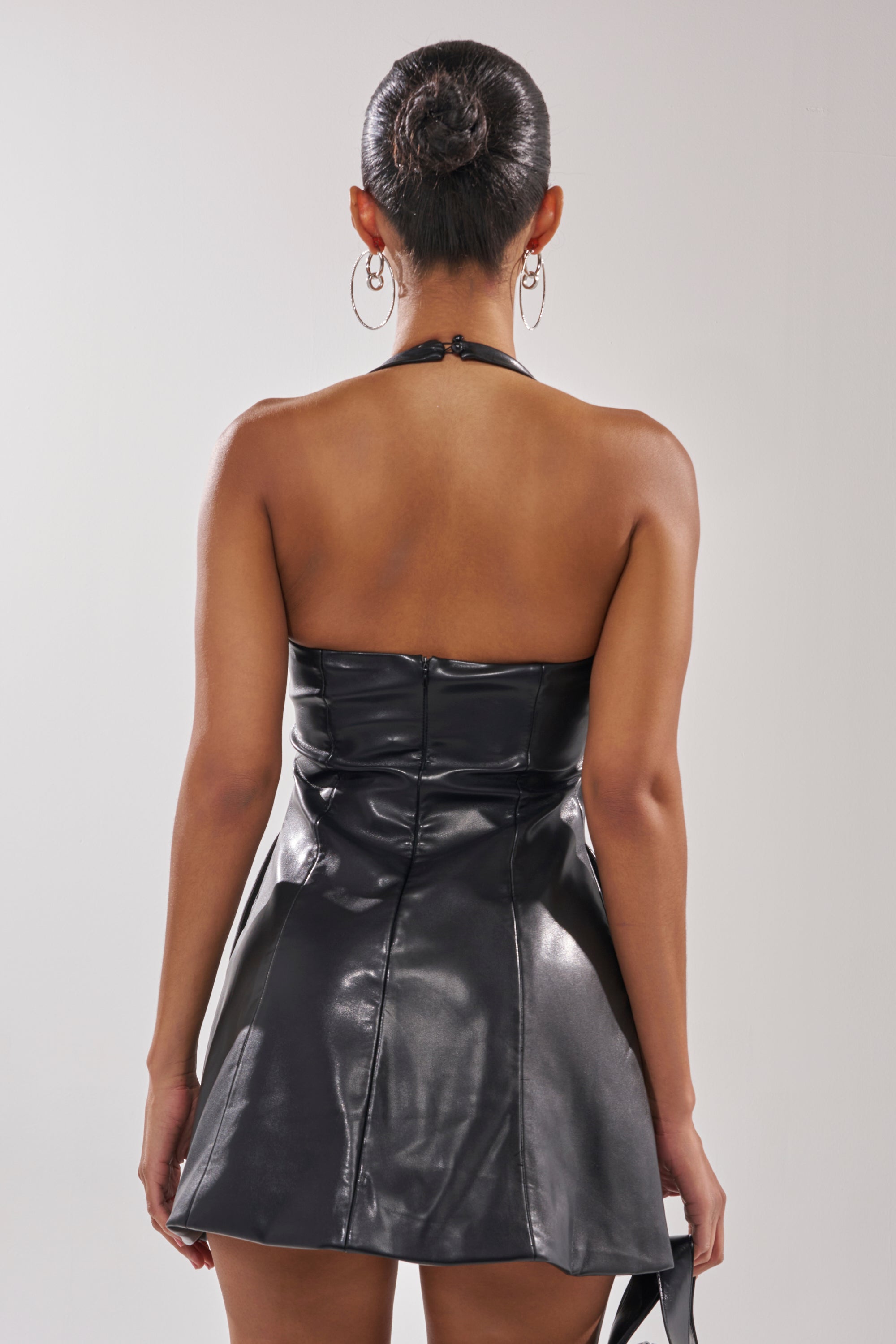 A woman is seen from behind wearing the ESSENTIAL HALTER NECK MINI DRESS in black faux leather. Her hair is styled in a neat bun, she sports large hoop earrings, and the background is plain and light-colored.