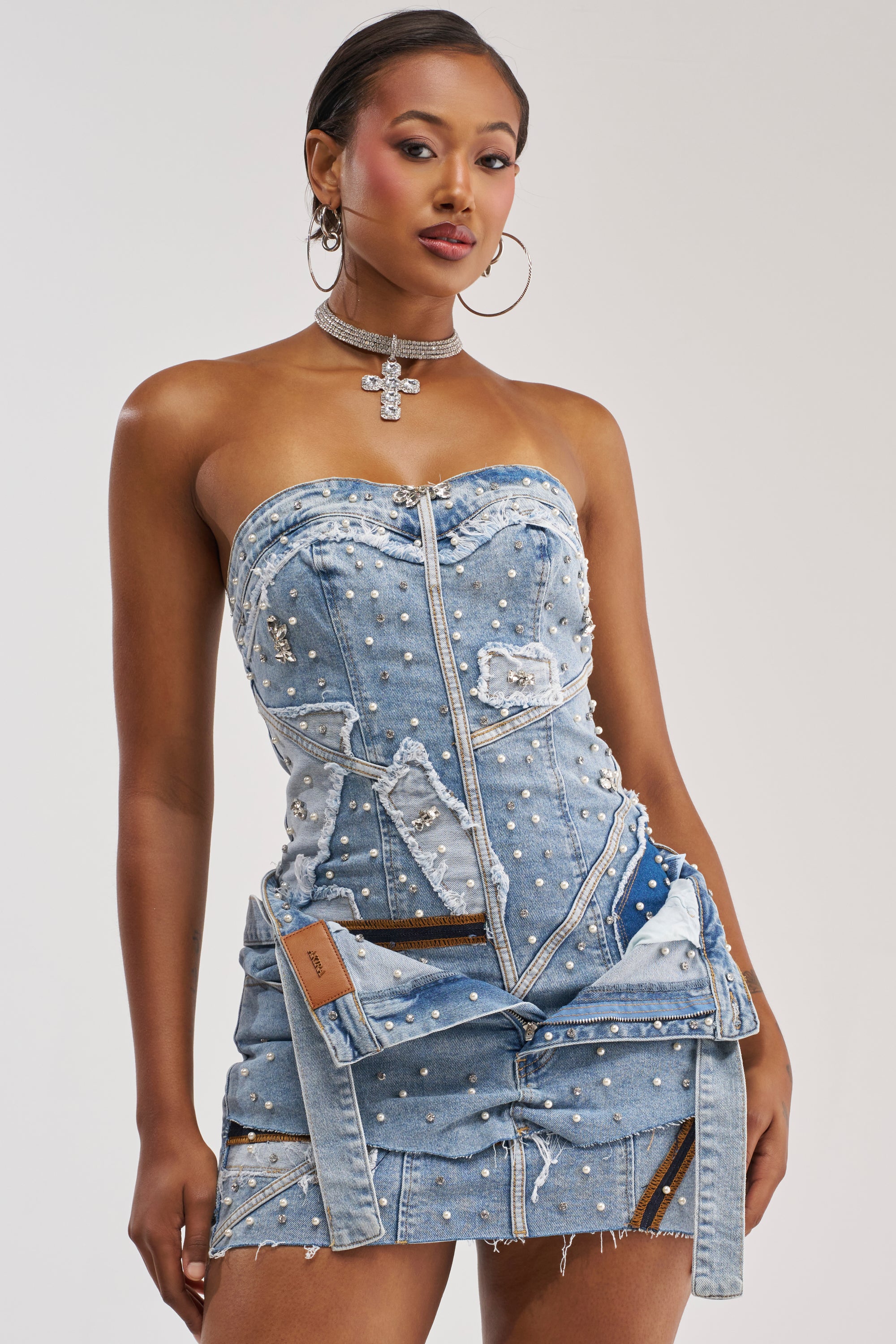 A woman wearing the THIS IS MY CALLING EMBELLISHED DENIM MINI DRESS—strapless, adorned with pearls and zippers—is accessorized with a cross choker necklace and hoop earrings as she stands against a plain background. This statement denim dress exudes edgy sophistication, making it the perfect festival outfit.