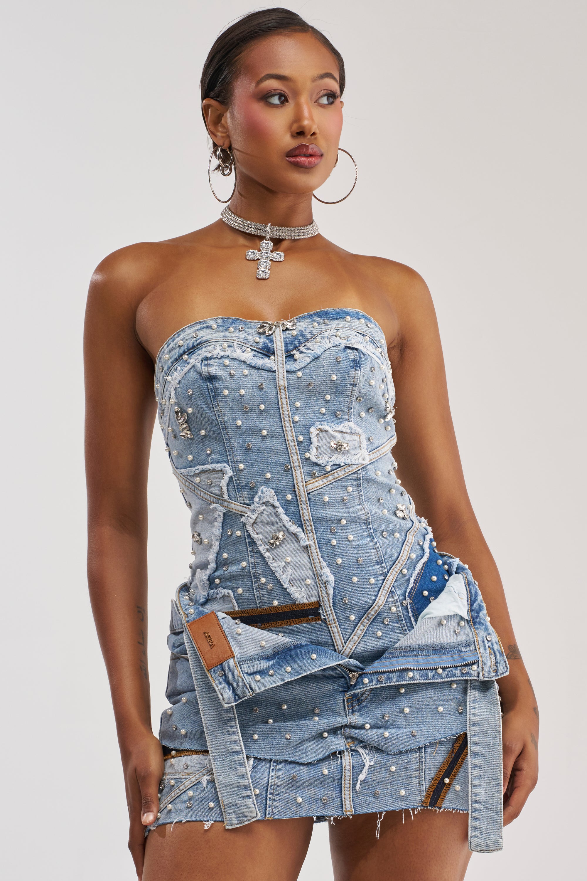 A woman poses against a plain, light background wearing the THIS IS MY CALLING EMBELLISHED DENIM MINI DRESS—a strapless denim mini dress adorned with pearls and zippers—styled with a rhinestone choker and hoop earrings for the perfect festival look.