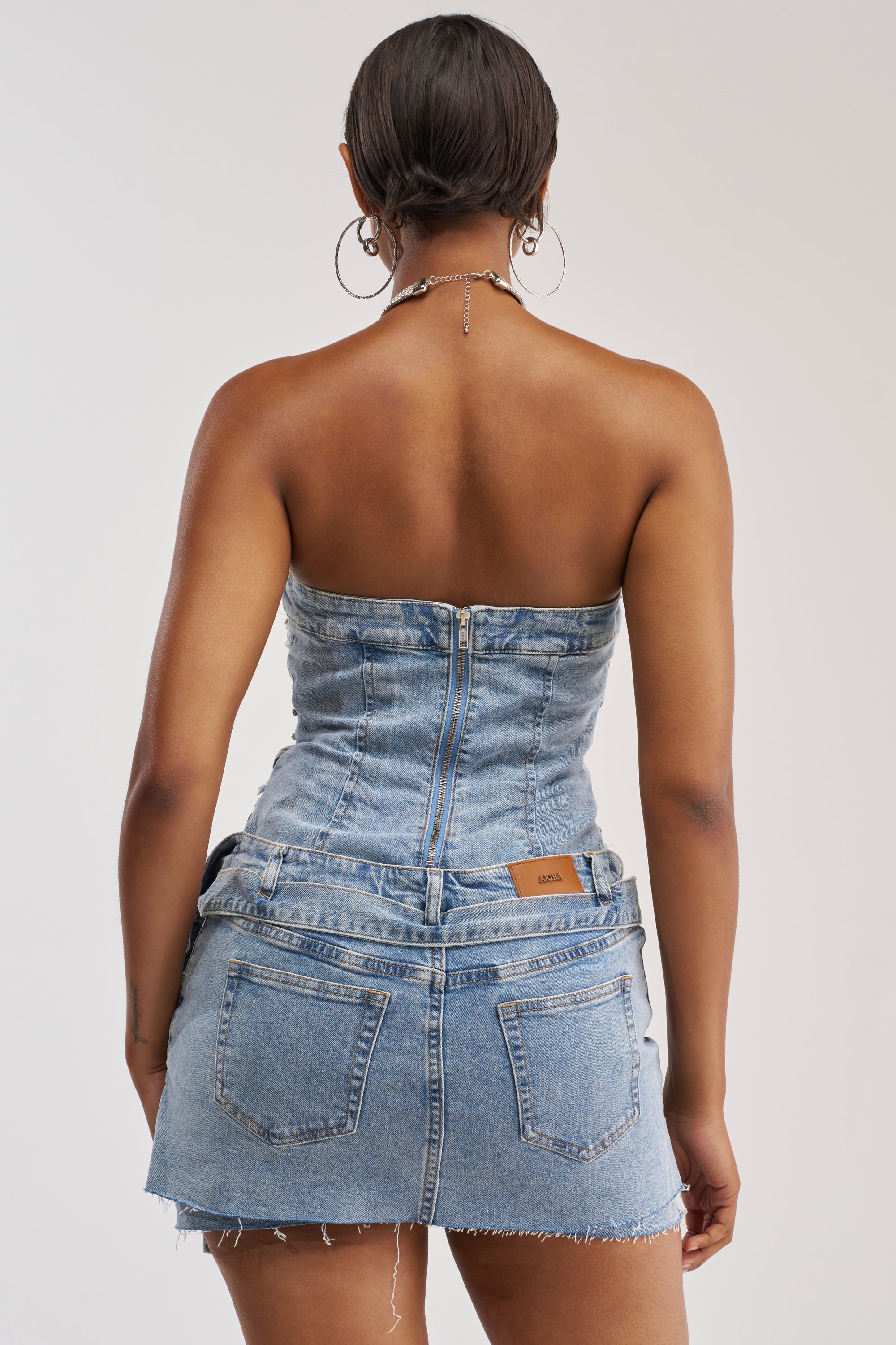 A person with short hair models the THIS IS MY CALLING EMBELLISHED DENIM MINI DRESS, a strapless mini dress with zipper accents, styled with large hoop earrings and photographed from the back against a plain backdrop—delivering an effortless festival-ready look in a minimalist setting.