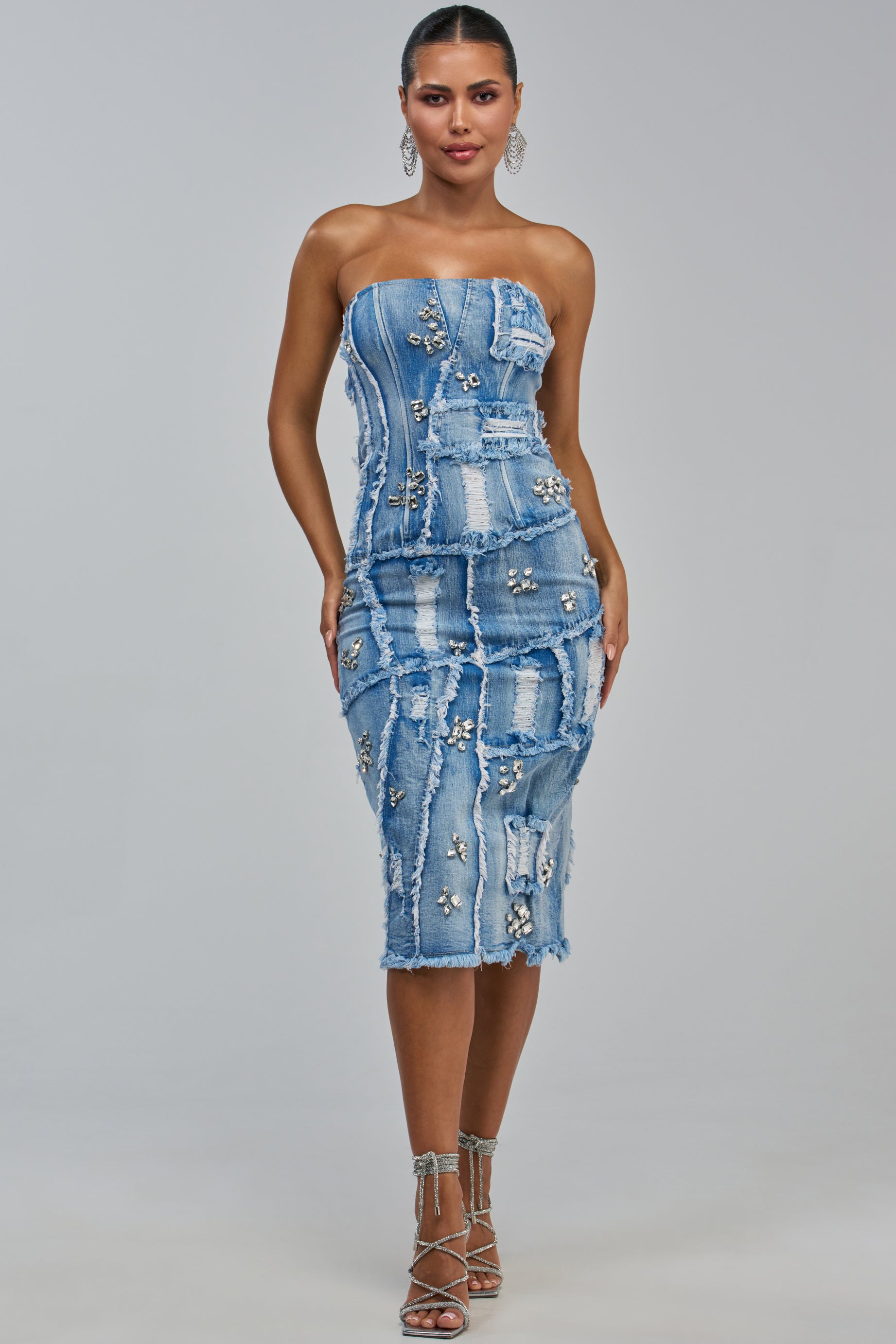 A woman stands facing forward against a plain light background, wearing the LELANI DISTRESSED RHINESTONE DENIM MIDI DRESS—a strapless, body-hugging dress featuring distressed denim, patchwork details, and shimmering rhinestone embellishments—paired with silver strappy heels.