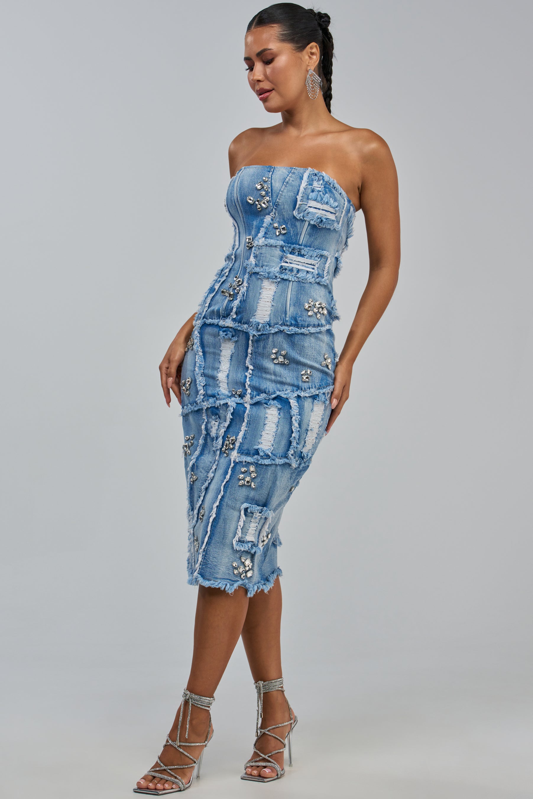 A woman models the LELANI DISTRESSED RHINESTONE DENIM MIDI DRESS—a strapless, fitted patchwork midi dress featuring distressed denim with frayed edges and rhinestone embellishments. She pairs it with strappy silver heels and stands against a plain, light grey background.