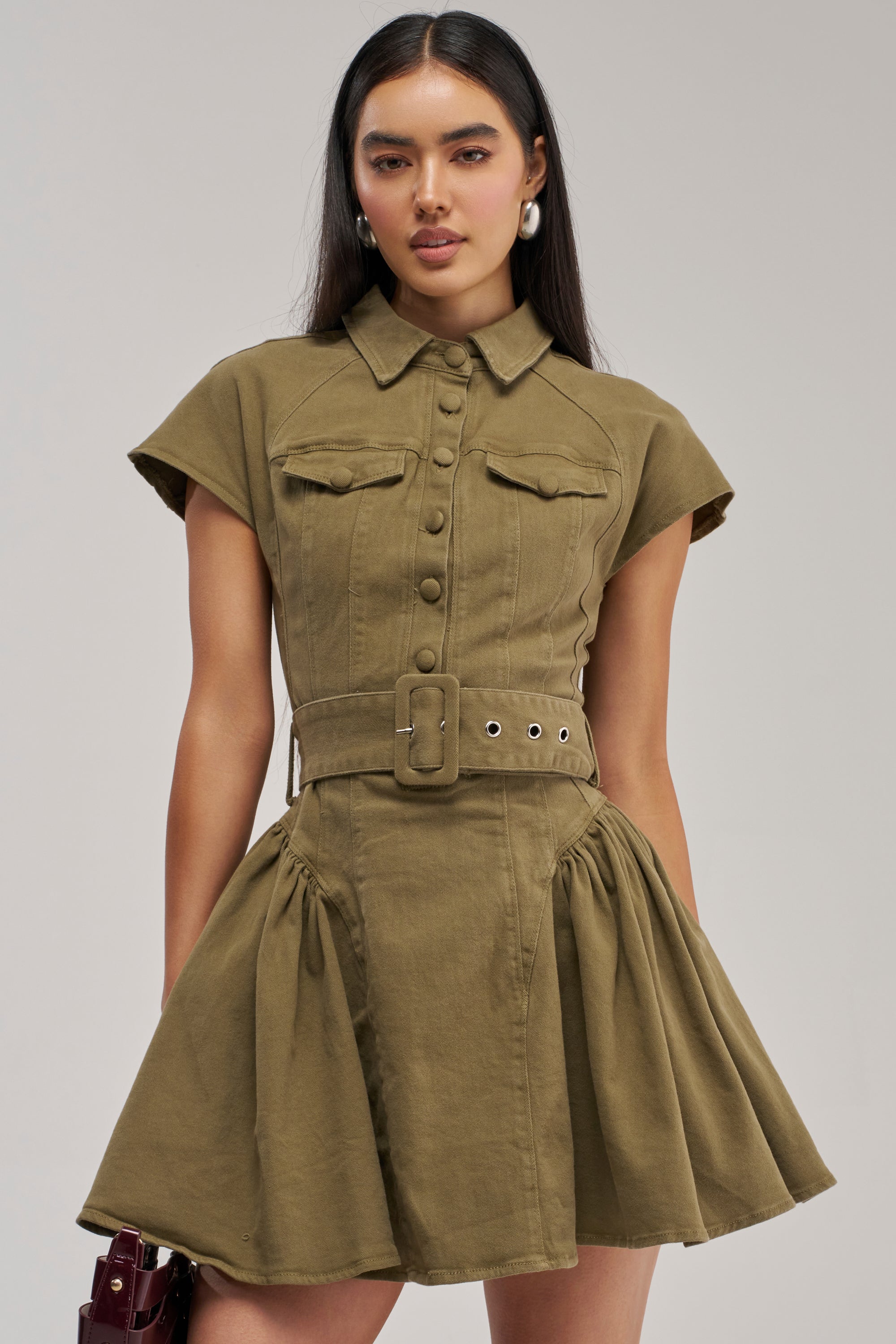 Wearing the SADIE BUTTON UP MINI DRESS IN OLIVE DENIM, this woman showcases a streetwear look with short sleeves, a belted waist, and a button-up front. The dress features a flared skirt and is styled simply with her long dark hair against a plain background.
