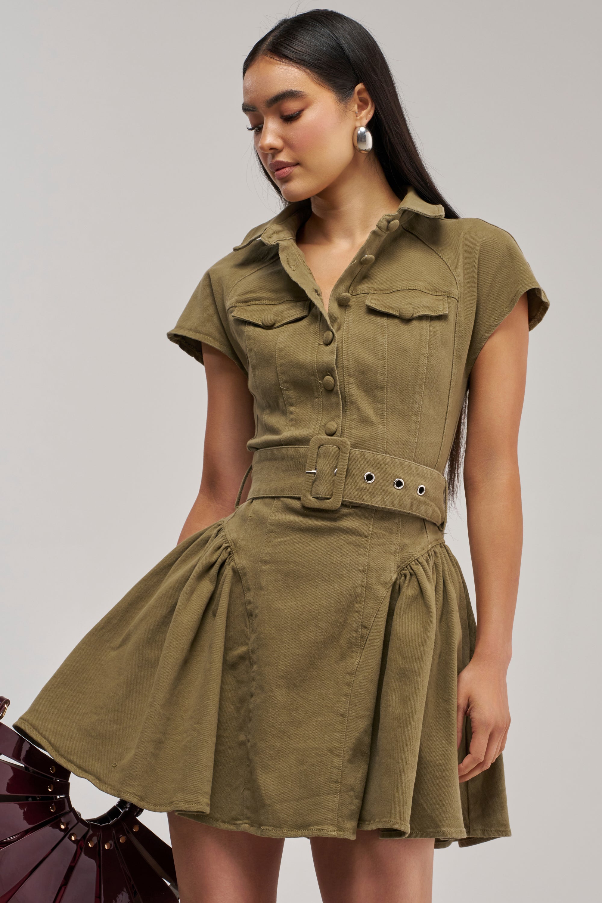 A woman stands against a plain background wearing the SADIE BUTTON UP MINI DRESS IN OLIVE DENIM, featuring short sleeves, a belted waist, chest pockets, a flared skirt, and silver hoop earrings. This streetwear dress effortlessly combines casual style with modern sophistication.